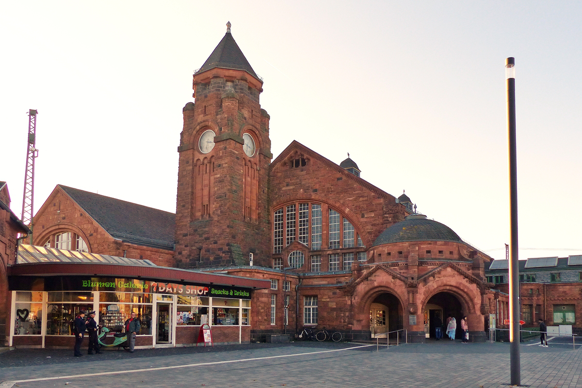 . Der Bahnhof von Gie�en von der Stra�enseite im Gegenlicht fotografiert am 01.11.2015. (Jeanny)

Bei dem Empfangsgeb�ude handelt es sich um ein dreigeschossiges, in rotem Sandstein gehaltenes Bauwerk mit einem markanten Uhrturm auf asymmetrischem Grundriss. Mit Planung und Ausf�hrung beauftragt war der Herborner Architekt Ludwig Hofmann. Das Empfangsgeb�ude schiebt sich als Keil zwischen die Main-Weser-Bahn und die in �stliche Richtung abzweigenden Gleise von der Vogelsbergbahn und der Strecke Gie�en–Gelnhausen.
