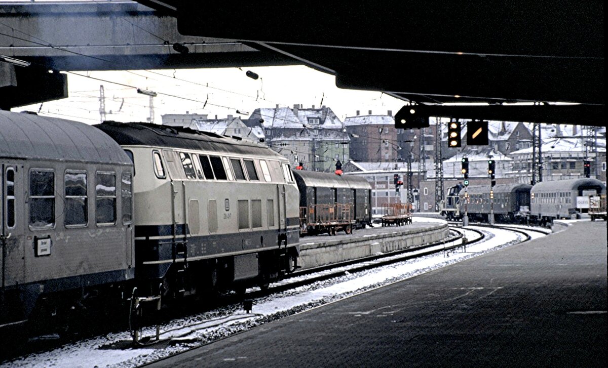 218 411-7 mit Nahverkehrszug fährt aus Um aus; vorn rechts rangiert eine 263, links steht ein Postwagen am Bahnsteig, am 15.10.1980.