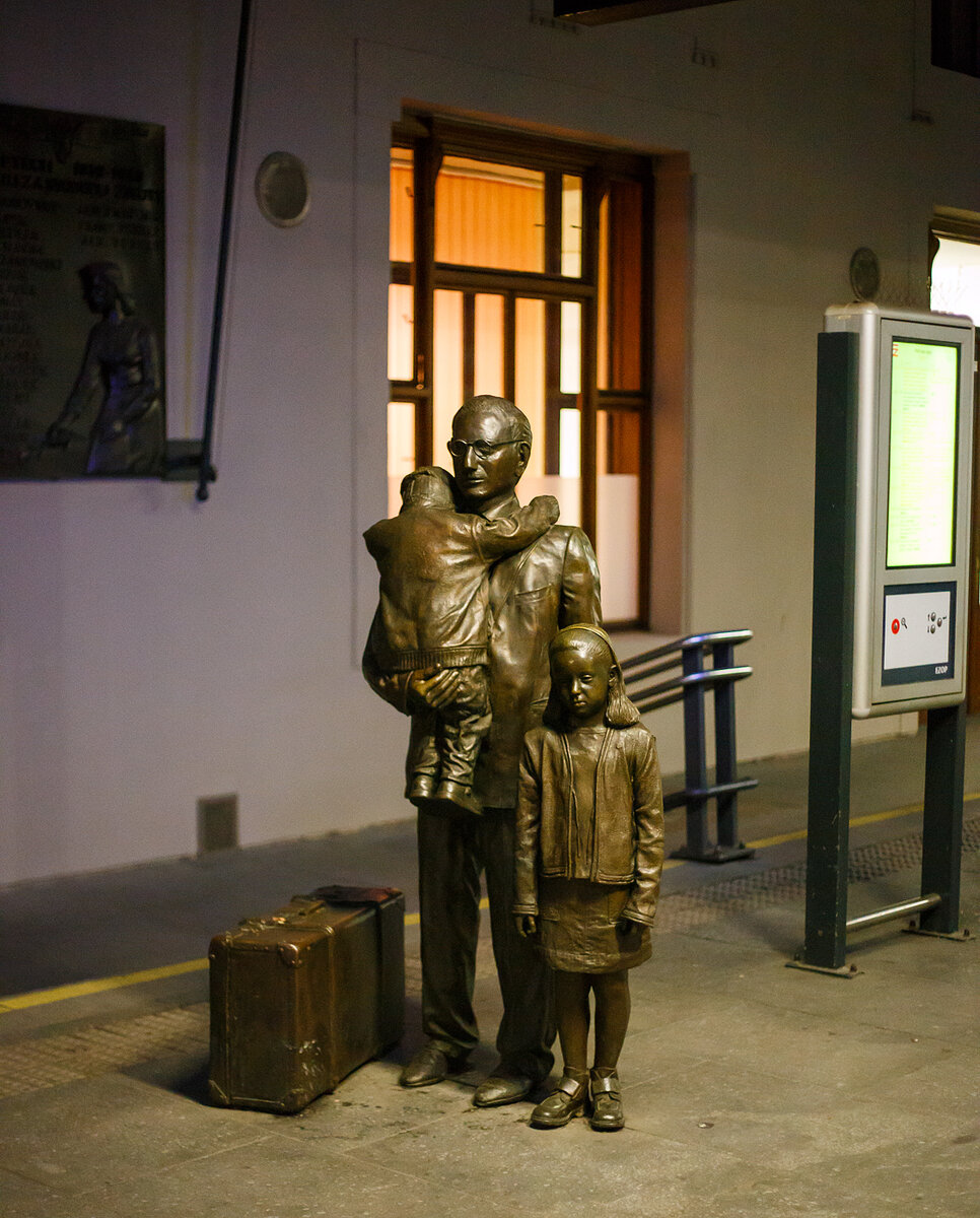 Bronzestatue einer tschechischen Familie am Bahnsteig 1 vom Prager Hauptbahnhof (Praha hlavni nadrazi) am 24.11.2022.  Die Skulpturengruppe ist von der Künstlerin Flor Kent. Ein Mann mit zwei Kinder und einem Koffer stehen in Bronze gegossen am Bahnsteig 1. Es sind sehr traurige Gesichter. Was ist die Geschichte dazu:

Das Denkmal ist sehr eindrücklich gestaltet. Die Personen stehen ohne Sockel quasi auf Augenhöhe mit den Wartenden am Bahnsteig 1 des Prager Hauptbahnhofs (Praha hlavní nádraží). Eine Tafel verrät Titel des Kunstwerks: Pro dítě (Für das Kind), Der Text dankt Sir Nicholas Winton (1909 – 2015), der kurz vor Beginn des Zweiten Weltkriegs die Rettung von 669 Kindern jüdischen Glaubens oder Herkunft aus der Tschechoslowakei vor dem Holocaust organisierte. Der Großteil davon waren Kinder aus Familien jüdischer Flüchtlinge aus Deutschland und Österreich, die in der Tschechoslowakei Asyl bekommen haben.
