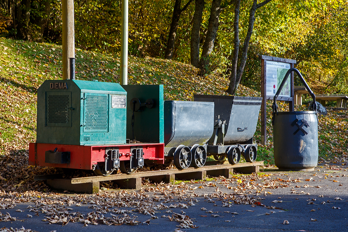 Die DIEMA Feldbahnlok vom Typ DL 6, Fabriknummer 1983, Baujahr 1956, Leistung 18 KW, Gewicht 1.115 kg, von 700 auf 600 mm um gespurt, als Denkmallok am 28.10.2021 mit zwei Grubenwagen beim Wodanstolln in Neunkirchen-Salchendorf. Wobei eigentlich ganz stilecht bzw. geschichtlich richtig ist dieses Denkmal nicht, denn die Lok ist eine Feldbahnlok. Bei den Siegerl�nder Erzgruben kamen aber Gubenloks zum Einsatz. 

Die DL 6 der Diepholzer Maschinenfabrik Fritz Sch�ttler (Diema) ist eine Diesellokomotive f�r Feldbahnen. Mit einem Gewicht von etwa 1 t, einer L�nge von ca. 1,7 m und einem Achsstand von 0,5 m geh�rt sie zu den kleinsten Lokomotiven �berhaupt. Die DL 6 entstand 1955 als kleinere und leistungsschw�chere Version der DL 8. Insgesamt wurden 220 Exemplare gebaut, womit die DL 6 zu den erfolgreichsten Diema-Konstruktionen geh�rt. Eingesetzt wurden die Lokomotiven vor allem von kleinen Torfabbaubetrieben. Die letzte Lokomotive wurde im September 1970 ausgeliefert.

Die Lokomotiven sind auf einem geschwei�ten Au�enrahmen aufgebaut. Die Breite des Rahmens hing von der Spurweite ab, insbesondere gab es eine Version f�r die in den Niederlanden verbreitete 700-mm-Spur, zu diese die Lok hier geh�rt. In der DL 6 kommen luftgek�hlte Einzylinder-Dieselmotoren zum Einsatz. Die Leistungs�bertragung erfolgt �ber einen Keilriemen vom Motor auf ein mechanisches Zweiganggetriebe und von dort aus �ber Rollenketten auf die beiden Achsen. Diese waren zun�chst �ber Evolutfedern abgest�tzt, sp�ter wurden Schraubenfedern eingebaut.

Der Fahrersitz ist, wie bei Lokomotiven dieser Kategorie �blich, quer zur Fahrtrichtung eingebaut, so dass der Fahrer seine Position bei Fahrtrichtungswechseln nicht �ndern muss. Motor und Getriebe nebst Schalthebeln befinden sich dabei rechts vom Fahrer, der Bremshebel links. Wobei bei dieser Lok der Fahrersitz fehlt.
