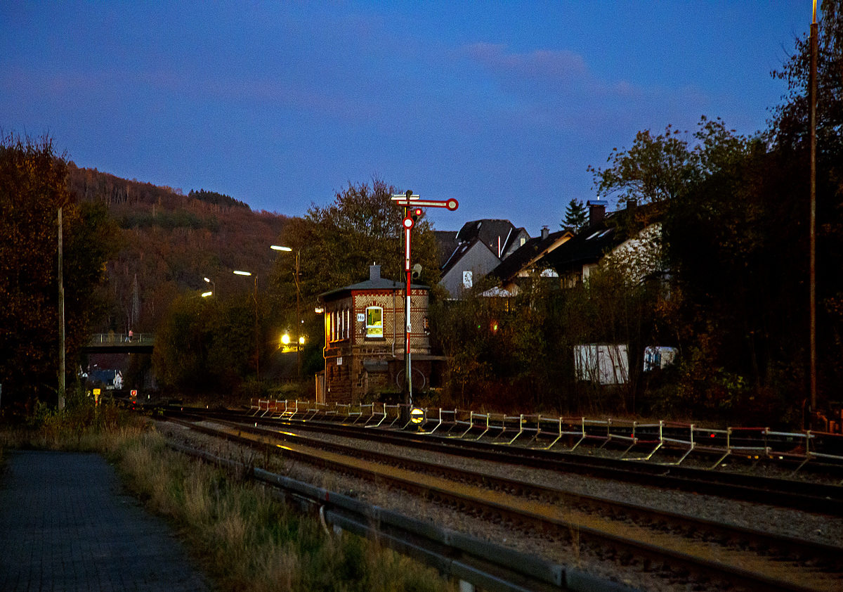 
Ein abendlicher Blick auf das 1901 gebaute Wärterhebelstellwerk Herdorf Ost (Ho) am 05.11.2020. 

Davor auf Gleis 2 das (Flügelsignal) Formhauptsignal, der obere Flügel zeigt waagerecht nach rechts und das obere Licht ist „rot“ und somit zeigt es Hp 0 (Halt) an. Unten vor dem Hauptsignal das Schutz- und Gleissperrsignal (waagerechter schwarzer Streifen auf runder weißer Scheibe), welches somit Sh 0 (Halt! Fahrverbot)  anzeigt.

Weit hinten kurz vor der Brücke sieht man das Signal Sh 2 – „Schutzhalt.“, die rechteckige rote Scheibe mit weißem Rand und davor das rote Licht. Eine Weiterfahrt ist somit auf der Hellertalbahn (KBS 462) in Richtung Neunkirchen nicht möglich, da noch Arbeiten ausgeführt werden.
