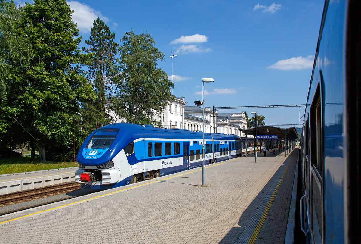 Ein letzter Blick aus dem Zug (Rx 760  Vy�ehrad ) heraus auf den Bahnhof Mari�nsk� L�zně (deutsch Marienbad) am 01.07.2015. 

Auf Gleis 2 steht der „RegioShark“  ČD 844 018-2 (CZ ČD 95 54 5 844 018-2), ein Dieseltriebzug vom Typ PESA LINK II, als Os 7382 zur Abfahrt bereit. Er hatte mich zuvor nach Marienbad gebracht.