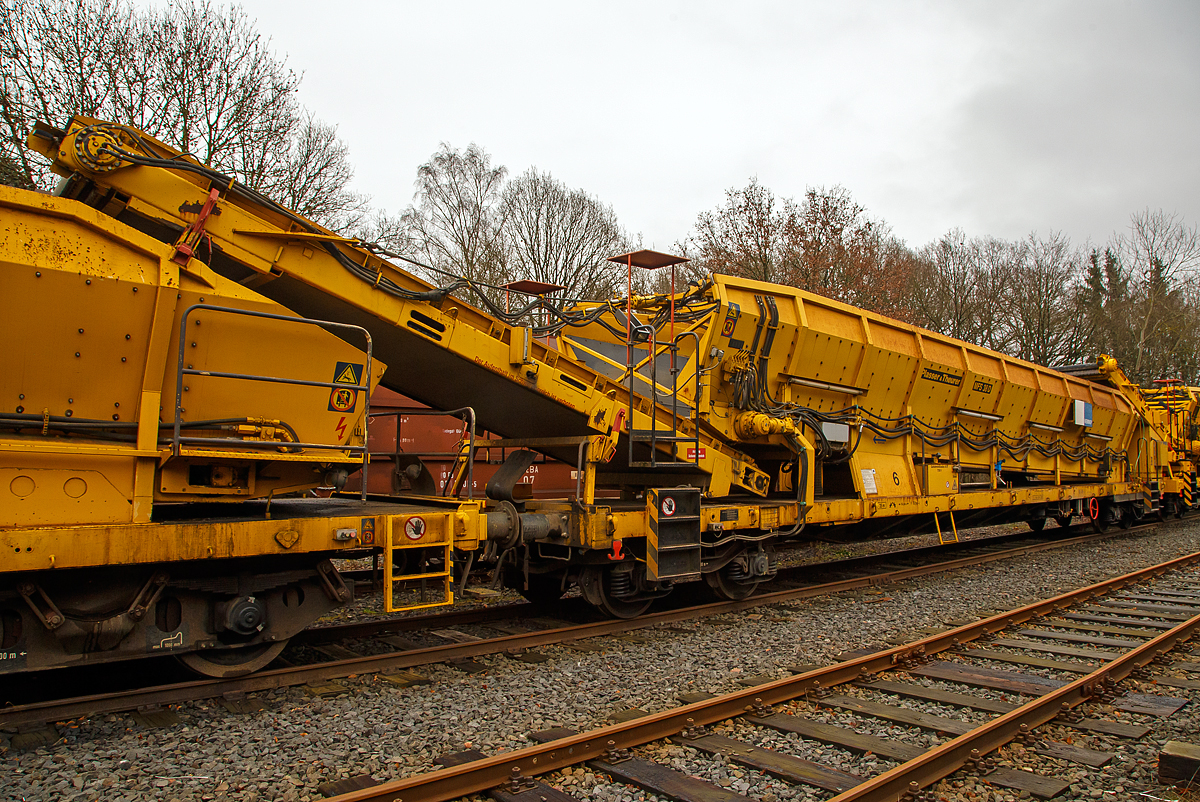 
Plasser & Theurer Material-, Förder- und Siloeinheiten MFS 38-D (teilweise auch BSW 2000 - Bunker-Schüttgut-Wagen genannt) der HERING Bahnbau, Burbach, Nr. 6 (Schweres Nebenfahrzeug Nr. D-HGUI 99 80 9552 125-3, ex 97 19 22 583 57-2, ist am 20.12.2020 auf dem Betriebshof der Westerwaldbahn (WEBA) auf der Bindweide bei Steinebach/Sieg abgestellt.

Diese MFS 38-D wurde 2002 von Plasser & Theurer gebaut. Die vierachsigen Fahrzeugrahmen und Drehgestelle sind moderne als die Wagen Nr. 1, 3 und 4 der Hering Bau.

Bei diesem MFS 38-D (das „D“ bedeutet Durchförderer) ist das Übergabeband starr (nicht schwenkbar), so ist ein  Entladen nebens Gleis nicht möglich. Die MSF 38-D haben ein Fassungsvermögen 38 m³ bzw. eine Nutzlast von 41,5 t, bei einem Eigengewicht von 38,5 t. Die Förderbänder werden hydraulisch angetrieben. Der Antrieb des Hydraulikaggregates erfolgt durch einen luftgekühlten Deutz Dieselmotor vom Typ F6L 912 mit einer Leistung von 82 kW.

TECHNISCHE DATEN:
Maschinenart: Material-Förder-Siloeinheit MSF 38
Hersteller: Plasser & Theurer
Spurweite: 1.435 mm (Normalspur)
Anzahl der Achsen: 4 (in 2 Drehgestellen)
Länge über Puffer:19.900 mm
Gesamtlänge mit Übergabeband: 23.550 mm
Drehzapfenabstand: 14.860 mm
Achsabstand im Drehgestell: 1.800 mm
Breite: 2.890 mm
Höhe: 4.280 mm
Eigengewicht: 38,5 t
Max. Zuladung (Nutzlast): 41,5 t
Ladevolumen: 38 m³
Höchstgeschwindigkeit: 100 km/h (geschleppt)
Kleister befahrbarer Gleisbogen: R 35 m
Bremse: KE – GP - A
Motor: luftgekühlter Deutz Dieselmotor vom Typ F6L 912 F
Leistung: 82 kW
Dieseltankvolumen: 750 l
Übergabeband: starr
Die Förderbänder sind in folgenden Geschwindigkeiten regelbar:
Siloband: 1. Gang (Speichern)   0 - 0,028 m/s / 2. Gang (Durchfördern)  0 - 0,140 m/s
Übergabeband: 0 - 1,000 m/s

Einsatz und Arbeitsweise:
Mit den Materialförder- und Siloeinheiten können unterschiedliche Schüttgüter wie beispielsweise Abraum, Planum- und Frostschutzmittel oder Bettungsstoffe unkompliziert, geladen, gefördert, gespeichert und entladen werden. Das Abraumverladesystem ist weitestgehend automatisiert und benötigt zur Bedienung nur wenig Personal. Das System verfügt zudem über eine autarke Energieversorgung (durch Dieselmotore). Ein unabhängiges Entladen ist durch schwenkbare Übergabebänder an jeder geeigneten Stelle möglich. Die MFS-Wagen können in Kombination mit einer Beladestation in beliebiger Anzahl aufgereiht und vor Kopf be- und entladen werden.

Am Boden jedes MFS 38 befindet sich ein über die ganze Wagenbreite gehender Fördergurt und an der Stirnseite ein breites Abgabeförderband. Das Aushubmaterial fällt von der Maschine kommend auf den Bodengurt, wird zum Abgabeförderband gebracht und an die nächste Einheit übergeben. Dieser Vorgang wiederholt sich, bis die am weitesten entfernte Siloeinheit erreicht wird. Bei dieser hintersten Siloeinheit ist das Abgabeförderband an der Stirnseite des Wagens in Ruhestellung. Auch der Fördergurt am Boden des Wagens ist so lange in Ruhestellung, bis die obere Füllgrenze erreicht ist. Dann wird der Fördergurt in Bewegung versetzt und dadurch im Übergabebereich freier Raum für die weitere Beladung geschaffen. Durch die dem Fülltempo angepasste Geschwindigkeit des Fördergurts wird die ganze Siloeinheit vollständig gefüllt. Das Fassungsvermögen einer Siloeinheit beträgt 38 Kubikmeter.

Nach Füllung einer Teilgruppe des Abraumzuges kann diese von der Restgruppe abgekuppelt werden, zu einer Entladestelle gefahren und dort entleert werden, während die Restgruppe des Abraumzuges noch befüllt wird. Das Entladeband ist nach beiden Seiten ausschwenkbar. Die Entladung kann dabei auf einer Deponie erfolgen oder es können LKW's oder Waggons am Nachbargleis beladen werden. Die MFS 38 (ohne nachfolgendes „D“) des Abraumzuges können durch Ausschwenken aller Abgabeförderbänder gleichzeitig entleert werden. Die Entladung eines Wagens dauert ungefähr 3,5 Minuten.
