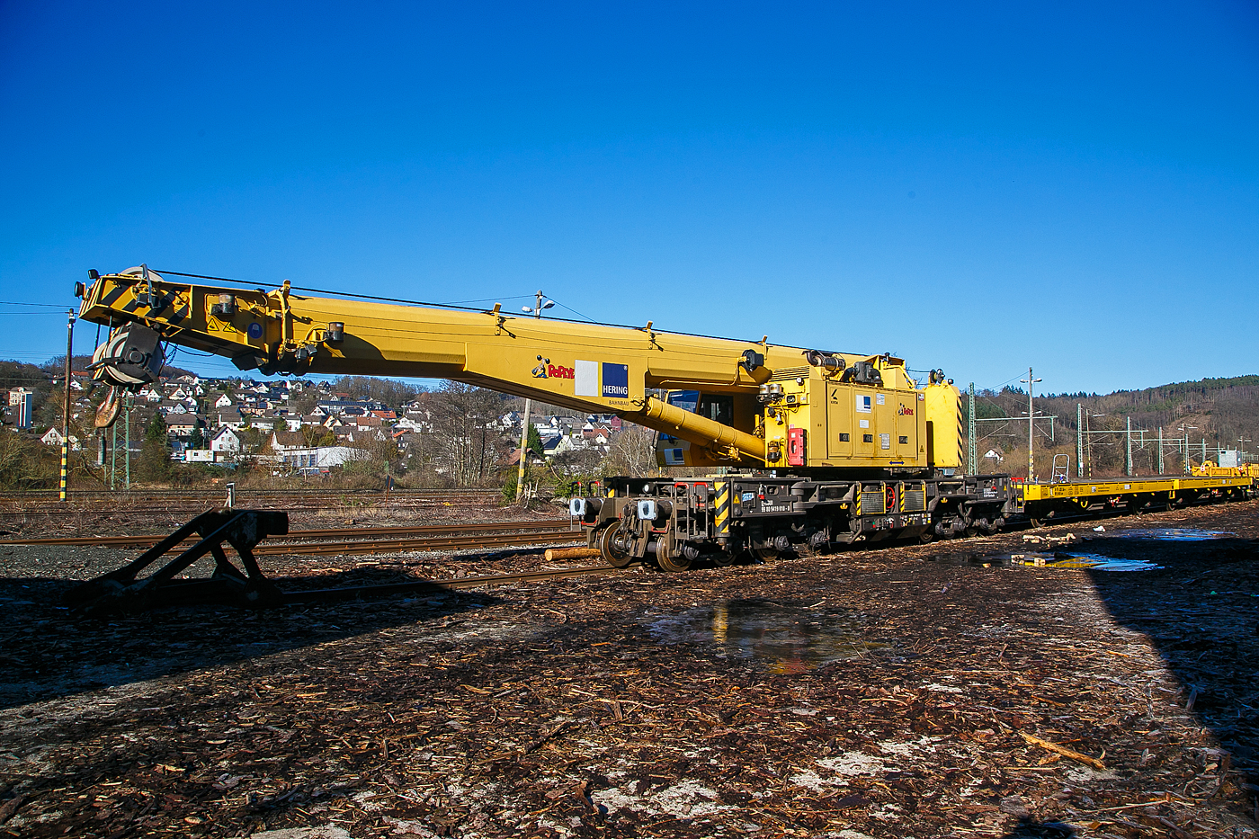 Beim Betriebshof der Westerwaldbahn (WEBA) auf der Bindweide bei Steinebach/Sieg hatte er neue Drehgestelle bekommen. Für die Fahrt hinauf, musste zu vor das Gegengewicht demontiert werden, damit Achslast gering genug war. Nun ist es aber wieder montier und er kann nun wieder in Einsatz gehen....
Der 125t - KIROW Gleisbauschienenkran KRC 810 T  Popeye  der Hering Bau (Burbach), Schweres Nebenfahrzeug Nr. D-HGUI 99 80 9419 010-0, steht am 08.02.2023 mit seinen Schutzwagen 24 80 4305 366-4 D-HGUI (Gattung Laas) und 33 80 3998 218-0D-HGUI (Gattung Res 072-1) beim Kleinbahnhof der WEBA in Scheuerfeld (Sieg).

Der Kran wurde 2013 von KIROW in Leipzig unter der Fabriknummer MT2012170324 gebaut und an die Hering Bau geliefert. Die Hering Bau gehört in Deutschland zu den größten Eisenbahnkran-Dienstleistern, sie hat 10 eigene Krane.

Der 125-t-Eisenbahndrehkran wird für den Aus-und Einbau von schweren Betonweichen, Gleisjochen und Hilfsbrücken sowie für die Montage von Ingenieurbauwerken eingesetzt. Bei Havarien auf Bahnstrecken leistet er schnelle Hilfe zum Bergen schwerer Lasten.

TECHNISCHE DATEN:
Länge über Puffer: 13.000 mm
Achsanzahl: 8
Drehzapfenabstand: 8.000 mm
Drehzapfenabstand im Drehgestell 1 und 2: 2.300 mm
Achsabstand in den Einzeldrehgestellen: 1.100mm
Ergebene Achsabstände in m: 1,1 / 1,2 / 1,1/ 4,6 / 1,1 / 1,2 / 1,1
Achsfolge: 1'A'1A'A1'A1'
Eigengewicht: 128.000 kg
Achslast: 16,0 t
Motorleistung: 235 kW
Fahrgeschwindigkeit im Zugverband: 100 km/h
Fahrgeschwindigkeit mit Eigenantrieb: 19 km/h
zul. Anhängelast: -
Zugelassen als Schwerwagen für Streckenklasse: CS 05
kleinster befahrbarer Radius: R 80 m

Tragfähigkeiten:
bei 0,00 m Ausladung vor Puffer, mit Schwerlastausrüstung: 125 t
bei 2,80 m Ausladung vor Puffer, ± 30°, abgestützt: 90 t
bei 2,80 m Ausladung vor Puffer, ± 20°, freistehend: 46 t

Übrigens: Bei einem KIROW KRC 910 wäre eine Demontage des Gegengewichtes nicht nötig, da dieser Krantyp in Transportstellung nur eine Achslast 13,0 t. Er verteilt ja auch die Lasten in Transportstellung über einen Gegengewichtwagen, sowie über den Ausleger-Schutzwagen. Gegengewicht und Ausleger werden auf diesen Wagen abgestützt.