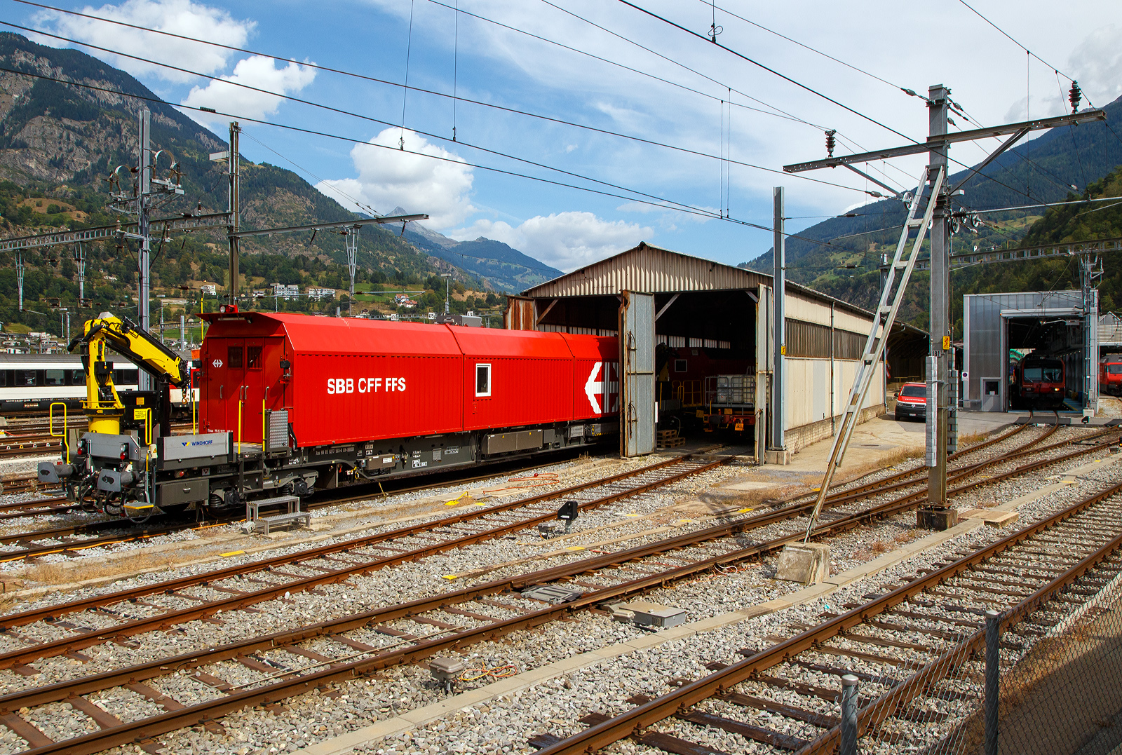 Blick aus einem in Brig einfahrenden MGB-Zug auf das SBB-Depot Brig am 07 September 2021, hier steht unteranderem der SBB Hilfswagen 2014 Xas 99 85 9277 503-0 CH-SBBI der SBB Infrastruktur. 

Zur�ck auf das Gleis:
Der Hilfswagen der SBB dient zur Bergung von entgleisten Eisenbahnfahrzeugen und ist der neuste seiner Generation. Er wird zum Einsatzort mit einer H�chstgeschwindigkeit bis zu 100 km/h geschleppt und ist mit einem Dieselpowerpack (6 Zylinder Dieselmotor von IVECO mit 260 kW Leistung) und einem hydrostatischen Antrieb ausger�stet. Der Hilfswagen ist mit zwei Drehgestellen ausger�stet, wobei eine Achse der beiden Drehgestelle als Antriebachse ausgef�hrt ist. Dies erm�glicht es, das Fahrzeug mit einer H�chstgeschwindigkeit von 7 km/h ohne Lokomotive zu verschieben. Der Aufbau des Fahrzeuges setzt sich aus zwei Ladekr�nen zum Heben von schweren Lasten, zwei Ger�tecontainern und einem Mannschaftscontainer zusammen.

TECHNISCHE DATEN Hilfswagen:
Allgemeine Daten:
Spurweite: 1.435 mm (Normalspur)
Achsfolge: 2´2´, am Einsatzort A1´ 2´-dh
L�nge �ber Puffer: 26.300 mm
Drehzapfenabstand: 19.000 mm
H�he der Ladefl�che (Containerauflagefl�che): 1.360 mm
Gewicht: 80.000 kg (Eigengewicht Wagen 55.000 kg)
Kleinster befahrbarer Gleisbogen: R 120 m

• Hersteller vom Basisfahrzeug und Arbeitsmodul: Windhoff Bahn- und Anlagentechnik GmbH
• Hersteller vom Lagersystem: Lista AG
• Hersteller Krane: Palfinger AG

Arbeitsmodul/Generator:
• Generator 96 kVA f�r Bordnetz (230 V, 400 V)
• Kranbahn im Fahrzeuginnern

Ausr�stung:
• Beleuchtungsmaterial
• Absperrmaterial
• Seilzugapparate
• Werkraum mit Werkbank und Werkzeugschr�nken
• Mannschaftsraum mit K�che
• Schneidbrenner
• Hilfskupplungen f�r Fahrzeuge mit automatischer Kupplung
• diverse Supportger�te
• Pioniermaterial
• elektrische und thermische Hydraulikaggregate
• diverse Hebezylinder (bis zu 120 Tonnen Hubkraft pro Zylinder)
• Aluminiumbalken und Panzerrollen f�r die horizontale Verschiebung von Schienenfahrzeugen
• diverses Holz f�r den Unterbau der Hebezylinder
• Hilfsdrehgestelle (Diplory)
• Krane Palfinger PK 8000 TC (2x) auf 4 Meter eine Hebekraft von 2 Tonnen und auf 7,5 Meter eine Hebekraft von 1 Tonne

Quelle: SBB Infrastruktur und Anschriften (eigene Sichtung)
