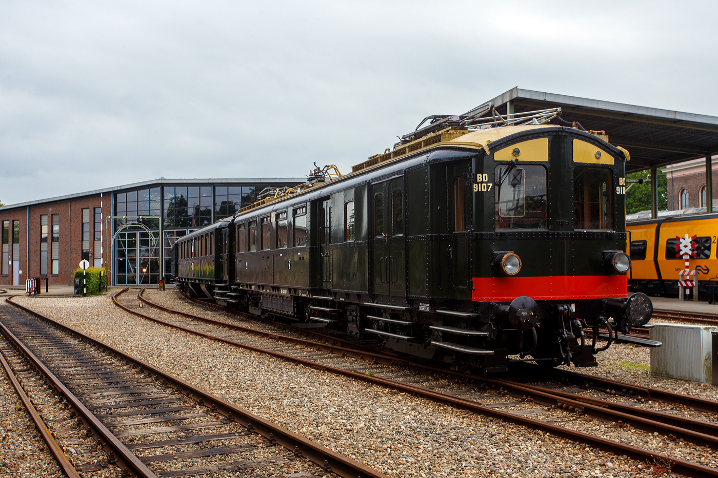 Der NS Mat ’24-Triebwagenzug (BD 9107, C 8553 und C 8104) „Blokkendozen“ (Kastendose) am 24 Juni 2025 im Het Spoorwegmuseum (bis 2005 NSM - Nederlands Spoorwegmuseum / Niederl�ndischen Eisenbahnmuseum) in Utrecht im ehemaligen Bahnhof Maliebaanstation. Ein Eisenbahnmuseum das zum Verweilen einl�dt. Der dreiteilige Zug vom Typ Mat’24 des Niederl�ndischen Eisenbahnmuseums besteht aus BD 9107, C 8553 und C 8104

Der Triebwagen BD 9107 (mBD):
Hier im Vordergrund der Triebwagen NS BD 9107 (mBD). Er wurde 1927 von Beijnes, offiziell Koninklijke Fabriek van Rijtuigen en Spoorwagens J.J. Beijnes (K�nigliche Fabrik f�r Kutschen und Eisenbahnwagen J.J. Beijnes) in Haarlem, mit einer elektrischen Ausr�stung von Metropolitan Vickers, gebaut und an die NS - Nederlandse Spoorwegen geliefert. Er hat 40 Sitzpl�tzen der zweiten Klasse sowie ein Gep�ckabteil. Seine Historischen Nummer waren ab 1927: BD 9007 / 1931: BD 9107 / 1946: CD 9107 / 1958: BD 5902 / 1966: 159016 / 1968: 9741937 / 1989 BD 9107.

Der Wagen kehrte Ende 1945 in besch�digtem Zustand aus Deutschland zur�ck. Er wurde 1947 instandgesetzt und als CD 9107 (mCD) wieder in Betrieb genommen. In den Jahren 1957/58 erfolgte der Umbau zu einem antriebslosen Wagen (BD 5902/BDz), dabei wurde die Inneneinrichtung der zweiten Klasse im „neuen Stil“ eingebaut, wobei die Anzahl von 40 Sitzpl�tzen erhalten blieb. Die elektrische Ausr�stung (einschlie�lich der Stromabnehmer) wurde entfernt, der F�hrerstand durch eine Plattform mit �bergangsfaltenbalg ersetzt und das Gep�ckabteil um einen Seitengang erweitert. Nur vier Jahre sp�ter wurde der Wagen �berfl�ssig und zur Nummer 159016, einem Schulungswagen f�r die Abteilung Gleisbau und Infrastruktur, umgebaut. Nach der Fertigstellung eines festen Schulungsraums im Jahr 1987 wurde der Wagen f�r die museale Erhaltung vorgesehen.

Die Restaurierung und der Umbau wieder zum Elektrotriebwagen BD 9107 erfolgten zwischen 1987 und 1989 im Ausbesserungswerk Amersfoort, dabei wurde das Fahrzeug weitgehend nach den Originalspezifikationen der 1930er Jahre hergerichtet, jedoch mit einer elektrischen Anlage der Firma Heemaf ausgestattet. Im Mai und Juni 2010 wurde der BD 9107 mit einer vereinfachten Version des automatischen Zugbeeinflussungssystems (ATBe) ausger�stet und im Strukton-Werk in Zutphen �berpr�ft. Die Betriebszulassung erfolgte im September, gefolgt von einer ersten Fahrt f�r die Ehrenamtlichen des Eisenbahnmuseums am 9. Oktober 2010. 

Im November 2011 wurden der BD 9107 und der Barwagen C 8553 zur �berholung der Fahrwerke in das NedTrain-Werk nach Haarlem �berf�hrt. Die eigentliche Aufarbeitung der Drehgestelle �bernahm die Firma Reuschling in Hattingen (Deutschland), dabei wurden unter anderem s�mtliche Tragfedern durch neue ersetzt. Im Dezember 2012 erhielt der gesamte Zug nach einer erfolgreichen Probefahrt im �ffentlichen Streckennetz die erneute Betriebszulassung. Der „Blokkendoos“-Zug ist aber derzeit aufgrund umfangreicher Instandhaltungsarbeiten au�er Betrieb.

Die NS-Baureihe Mat ’24 oder Materieel ’24 war eine (die erste) Serie von Elektrotriebz�gen, die von der NS - Nederlandse Spoorwegen (Niederl�ndischen Staatsbahnen, heute Niederl�ndische Eisenbahnen AG) ab 1924 in gro�er Zahl eingesetzt wurde. Diese Z�ge sind unglaublich robust und schwer. nd der Tatsache, dass die Trainer wie Bausteine zusammengestellt werden k�nnen. Im Volksmund erhielten sie wegen des eckigen �u�eren und der Tatsache, dass die Wagen wie Bausteine zusammengestellt werden konnten, den Spitznamen „Blokkendozen“ (Kastendose). Ein weiterer Spitzname war „buffermaterieel“, weil sie im Unterschied zu anderen, mehr windschl�pfig aussehenden elektrischen Triebz�gen mit �blicher Schraubenkupplung versehen waren. Wegen der Verbindung �ber Schraubenkupplung mit zwei Puffern konnten die Einheiten sehr flexibel kombiniert werden. Meist bestand ein Zug aus zwei Triebwagen und zwei oder drei antriebslosen Mittelwagen. Die lauten Ger�usche der Motoren und Antriebe erinnerten manche Fahrg�ste an Staubsauger, so dass auch dieser weitere Spitzname  Staubsauger  verwendet wurde. 

Ab Mitte der 1950er Jahre wurden die Mat ’24 zunehmend durch die neue Baureihe Mat ’54 ersetzt. Der letzte elektrische „Blokkendoos“-Zug verkehrte am 31. Mai 1959. Einige Wagen blieben als Reisezugwagen bis 1973 im Einsatz, sie wurden unter anderem in Milit�rz�gen sowie zur Kapazit�tserweiterung w�hrend der Hauptverkehrszeiten verwendet. Bei einigen Triebwagen wurden die Stromabnehmer und Motoren demontiert, sie wurden auch als einfache Beiwagen weiterverwendet. Da die Mat ’24 sehr robust waren, wurden einige Einheiten sp�ter auch als Postwagen oder f�r Dienstzwecke eingesetzt. Mehrere Wagen blieben museal erhalten.

TECHNISCHE DATEN Triebwagen BD 9107 (mBD):
Baujahr: 1927
Spurweite: 1.435 mm (Normalspur)
L�nge �ber die Puffer:	19.800 mm
Achsfolge:	Bo'Bo'
Achsabstand im Drehgestell: 2.500 mm
Treibraddurchmesser: 1.000 mm (neu)
Eigengewicht: 59.000 kg
Tragf�higkeit Gep�ckraum: 2,5 t
Traktionsmotoren: 4 St�ck vom Typ Heemaf TM 8
Stundenleistung: 4 x 136 = 544 kW (Urspr�ngliche Leistung 661,5 kW)
Dauerleistung: 4 x 100 = 400 kW
�bersetzung: 27 : 56
H�chstgeschwindigkeit: 100 km/h
Kupplung: Schraubenkupplung (UIC-Kupplung)

