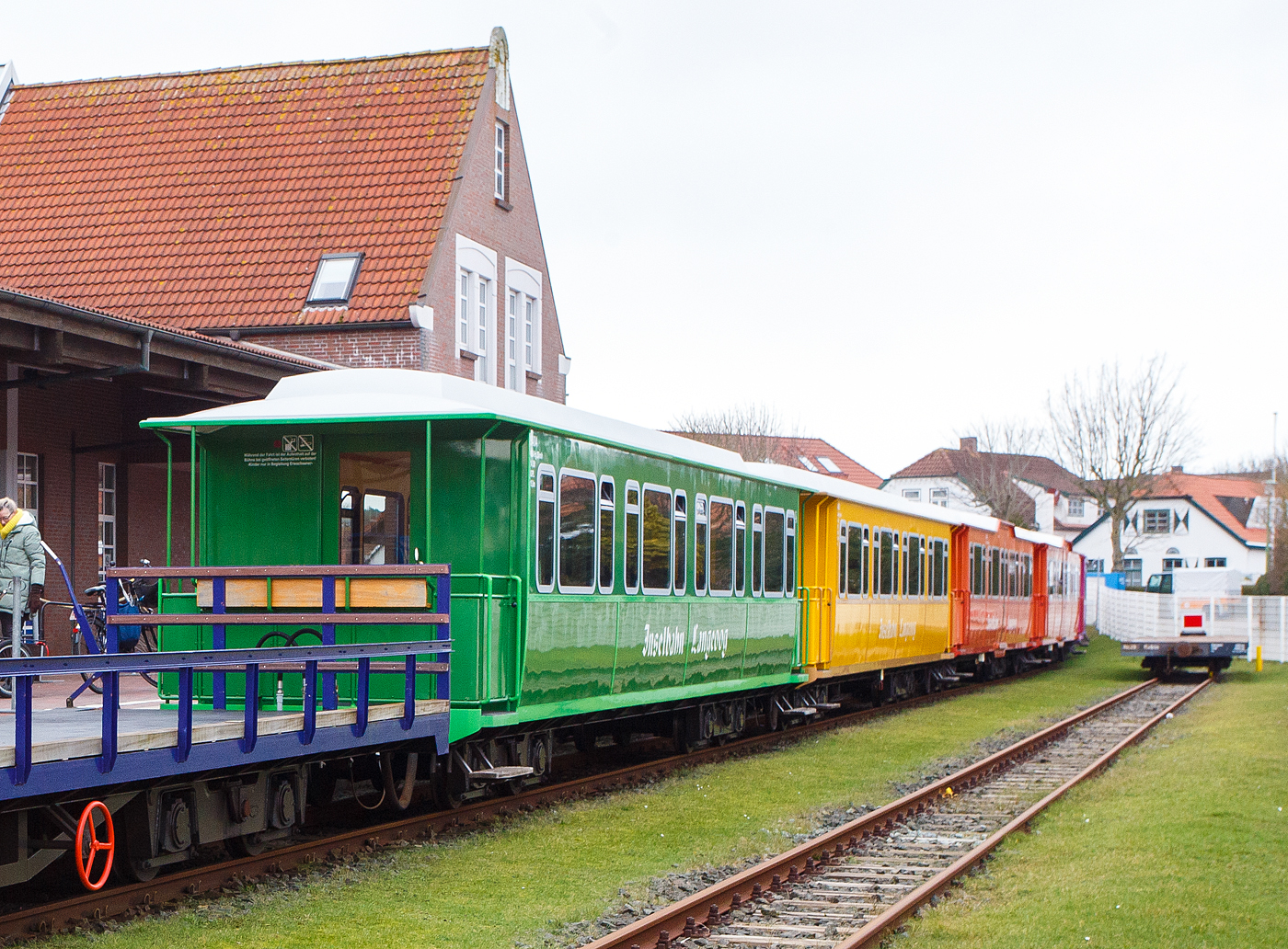 Der vierachsige meterspurige Personenwagen 94/04 (Sitzwagen) der Gattung KB4 der Inselbahn Langeoog vom Eigenbetrieb der Schiffahrt der Inselgemeinde Langeoog am 13 Februar 2024 im Insel Bahnhof Langeoog (im Ort Langeoog).

Der Wagen wurde von der Bremer Waggonbau GmbH 1994/95  gebaut. Die Serie für Inselbahn Langeoog bestand aus 10 Personen- und 1 Containertragwagen. Mit diesen elf neu gebauten Wagen für die Inselbahn Langeoog endete die Geschichte der Bremer Waggonbau GmbH, sie ging anschließend in Insolvenz, aber nicht wegen der Inselbahn. 

TECHNISCHE DATEN der Sitzwagen 94/01 bis 94/08:
Spurweite: 1.000 mm
Achsanzahl: 4
Länge über Kupplung: 15.000 mm
Länge über Rahmen: 14.200 mm
Drehzapfenanstand: 8.970 mm
Achsabstand im Drehgestell: 1.800 mm
Laufraddurchmesser: 650mm (neu)
Breite: 2.550 mm
Höchstgeschwindigkeit: 25 km/h
Eigengewicht:  14.500 kg
Sitzplätze: 62

Für interessierte im Anhang eine etwas längere Beschreibung der Wagen:
Die Fahrzeugrahmen bestehen aus handelsüblichen U- und Winkelprofilen der Stahlgüte St 52-3, die miteinander verschweißt wurden. Die Wagenkästen, die mit Edelstahlblechen außen verkleidet sind, wurden nicht als Ganzes auf den Fahrzeugrahmen aufgesetzt, sondern entstanden in einzelnen Segmenten, die dann auf den Rahmen aufgesetzt und angepasst wurden, dann wurden sie auf diesen verschweißt. Zusammen mit dem ebenfalls aus Edelstahl bestehenden Dach bilden Fahrzeugrahmen und Aufbau eine sehr verwindungssteife Einheit. Der Grund der Verwendung von Edelstahl ist die salzhaltige Luft an der Nordsee, normaler Baustahl oxidierend (rostet) hier viel schneller. Bevor die ersten Lackierarbeiten ausgeführt wurden, sind die auch aus Edelstahl bestehenden Bühnengeländer montiert worden, die gleichzeitig als Regenfallrohre für das Wagendach dienen. Die Edelstahlbleche des Wagenkastens wurden von innen und außen gesandstrahlt,  um eine bessere Haftung für den Farbaufbau zu erreichen.

Besonderer Wert wurde auf den Korrosionsschutz der Wagen gelegt, damit sie möglichst lange dem rauen Nordseeklima widerstehen können, Zur Minderung des Körperschalls wurde im Wageninnern Antidröhn-Masse aufgetragen und der Wagen mit Isolierungsmaterial ausgekleidet. Der Fußboden ist zweischalig ausgeführt und kann deshalb einen breiten Kabelkanal aufnehmen, in dem alle wichtigen Kabelverbindungen zusammengefasst sind. Die elektrische Ausrüstung der Wagen lieferte und montierte die Fa, Pintsch-Bamag, Alle Wagen sind mit einer elektrischen Heizung, einer Lautsprecheranlage und einer elektrischen Beleuchtung ausgestattet, Die beiden Mehrzweckwagen verfügen zudem über elektrisch ausfahrbare Laderampen für Rollstuhlfahrer, Kinderwagen usw.. Solange die Rampe ausgefahren ist, läßt sich aus Sicherheitsgründen der Zug nicht in Bewegung setzen. Für den Lokführer sind entsprechende Warnlampen auf dem Bedienungspult der Lok angebracht.

Nach dem Lackieren der Wagen mit einem seewasserbeständigen Farbsystemen und drr zuvor erfolgten Montage den genieteten Flacheisen (als Verzierung) außen am Wagenkasten wurden die isolierverglasten, getönten und druckdichten Fenster eingebaut. Die zahlreichen Abdeckleisten und die Holzlattensitzbänke wurden aus Eschenholz gefertigt und klarlackiert. Der Wagenboden ist mit einem bräunlich marmorierten PVC-Bodenbelag ausgestattet.

Eine technische Besonderheit stellen die neuentwickelten Drehgestelle dar. Ihre Rahmen wurden aus einzelnen vorgeformten Blechteilen zu einem verwindungssteifen Hohlkasten verschweißt. Aufgrund der örtlichen Gegebenheit war eine besonders niedrige Drehgestellbauart erforderlich, außerdem wurde großer Wert auf einen möglichst geringen Wartungsaufwand gelegt, Die Radsätze sind deshalb mit Megi-Federn im Drehgestellrahmen abgefedert. Einen geringen Wartungsaufwand verspricht man sich auch von den vom Lkw-Anhängerbau bekannten Drehkränzen, die anstelle der im Eisenbahnfahrzeugbau üblichen Drehzapfen mit seitlicher Wagenkastenabstützung zum Einbau gelangt sind. Der Drehkranz ist fest mit dem Fahrzeugrahmen verschraubt und über eine Traverse gelenkig auf dem Drehgestell befestigt. Die gelenkige Traverse, war erst nicht vorgesehen, aber man musste feststellen das man sie für eine sichere Spurführung braucht.

Alle 11 Wagen sind mit einer Feststeilbremse und 5 mit einer zusätzlichen Druckluftbremse ausgerüstet (Wagen 94/06 bis 10), Dabei ist jeweils nur ein Radsatz und nur in einem Drehgestell pro Wagen über eine Scheibenbremse abgebremst, Das so erzielte Bremsgewicht von 7 t reicht unter den betrieblichen und topographischen Verhältnissen der Inselbahn wohl aus. Die Feststeilbremse wird mit einem Handrad betätigt, das auf einer der offenen Bühnen montiert ist. Mit dem Drehen des Handrades wird aber kein Bremsgestänge in Bewegung gesetzt, sondern ein ausgeklügelter Bowdenzug-Mechanismus betätigt direkt die Bremszange an der Bremsscheibe.

Die Radsätze haben einen Laufkreisdurchmesser von 650 mm und sind mit Kegelrollenlagern ausgestattet, Die Radsätze wurden von SCHÖMA komplett gefertigt, mit montierten Achslagern und an die Bremer Waggonbau geliefert und dort in die vorbereiteten Drehgestellrahmen eingebaut.

Auch wenn nicht alle Wagen fertig waren, ließ die Inselbahn Langeoog Anfang Februar 1995 alles aus Bremen abtransportieren was mit ihren neuen angelieferten Loks zu tun hatte. Aber auch der Konkurs der Bremer Waggonbau GmbH zeichnete sich wohl schon stärker ab, als es mancher wahrhaben wollte. Den wenn die Insolvenz angemeldet ist kann keiner mehr einfach seine Ware abholen. Daher gelangten die beiden Mehrzweck- und der Containertragwagen unvollendet nach Langeoog.

Alle 10 Wagen wurden der Urlaubsstimmung der Fahrgäste entsprechend farbenfroh gestaltet. 

Für Fachleute nachfolgend Aufstellung der jeweiligen Wagenkasten-Grundfarben sowie der Zierlinien und der Rahmen mit Angabe der RAL –Nummern, sie kann einen Eindruck vom Erscheinungsbild der Zuggarnitur vermitteln:
94/01: Himmelblau/Enzianbiau (5015/5010),
94/02: Enzianblau/Himme!biau (5010/5015),
94/03: Chromgelb/Gelborange (1007/2000),
94/04: Gelbgrün/Grasgrün (6018/6010),
94/05: Grasgrün/Gelbgrün (6010/6018),
94/06: Kadmiumgelb/Braunbeige (1021/1011),
94/07: Verkehrspurpur/Blaulila (4006/4005),
94/08: Kadmiumgelb/Braunbeige (1021/1011),
94/09: Blutorange/Tomatenrot (2002/3013).
94/10: Blutorange/Tomatenrot (2002/3013),
94/11: Orange (RAL 2000)
