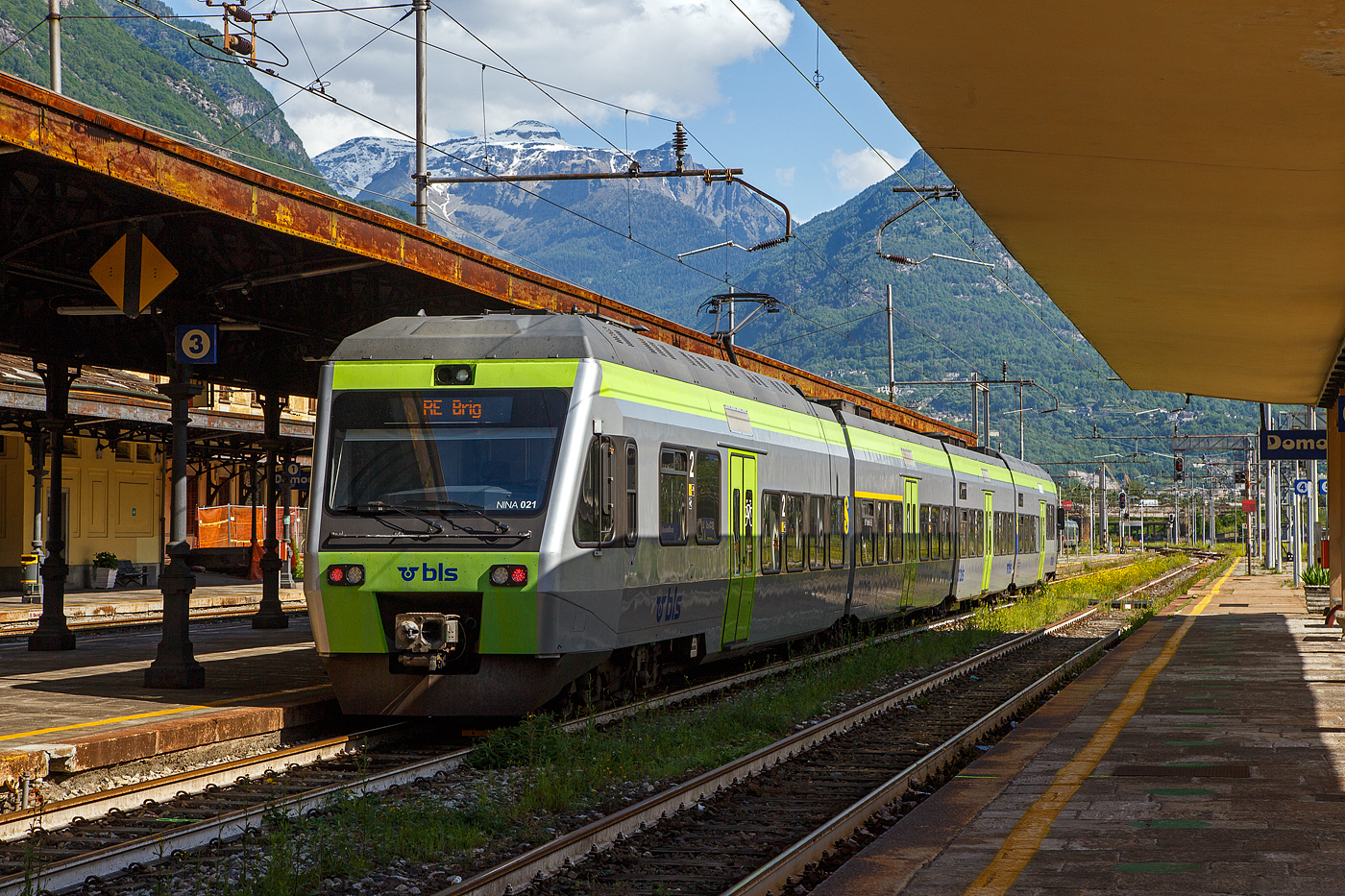 Der vierteilige BLS NINA 021 - RABe 525 021(94 85 7525 021-2 CH-BLS) steht am 26. Mai 2023 als RE nach Brig im Bahnhof Domodossola zur Abfahrt bereit.

Eigentlich sollten die BLS RABe 528 “MIKA” von Bern �ber die L�tschberg-Bergstrecke (Kandersteg) und Brig nach Domodossola fahren, aber diese haben noch keine Zulassung f�r Italien, die sie hinter dem SBB) Simplontunnel bis Domodossola brauchen. Und so wird der Zuglauf in Brig gebrochen und “NINA” fahren (min. bis zum Sommer) als Ersatz.

Von den BLS �NINA� (Niederflur-Nahverkehrszug) wurden 41 St�ck zwischen 1998–2005 in 5 Serien von Vevey Technologies SA in Villeneuve (vormals ACMV - Ateliers de constructions m�caniques de Vevey) ab 2001Bombardier Transportation zusammen mit Bombardier-Talbot in Aachen (D)gebaut, die elektrische Ausr�stung wurde von Alstom/Traxis (vormals Holec) zugeliefert. An die zweite Bestellung der BLS schlossen sich auch die Transports de Martigny et R�gions (TMR) und die Transports R�gionaux Neuch�telois (TRN) an, diese werden jedoch als RABe 527 gef�hrt. 

Die klimatisierten Niederflur-Nahverkehrsz�ge „NINA“ bieten modernen Fahrkomfort in erfrischendem Design und verkehren sowohl als 3-teilig wie auch als 4-teilige Komposition. Tiefe Einstiege und gro�e Plattformen sorgen f�r Bewegungsfreiheit, und Geschwindigkeiten von bis zu 140 km/h garantieren attraktive Fahrzeiten. Die dreiteiligen bieten 
148 und die vierteiligen 191 Sitzpl�tzen. Es k�nnen bis zu 4 Triebz�ge aneinandergekoppelt werden (maximal 592 bis 764 Sitzpl�tze).

Das Fahrzeug wurde Mitte der 1990er-Jahre von Vevey Technologies zusammen mit Bombardier-Talbot in Aachen (D), entwickelt und gebaut. Das Niederflurkonzept und die Drehgestell-Technik stammen weitgehend vom Triebzug Talent, der von Bombardier-Talbot in Aachen entwickelt wurde. Die Drehgestelle weisen eine fast gleiche Konstruktion auf, die lediglich f�r die NINA angepasst wurde. F�r die Elektrotechnik war die Firma Holec Machines & Apparaten – die sp�tere Alstom/Traxis – aus Ridderkerk (NL) zust�ndig, die, wie die Vevey Technologies zu dieser Zeit auch, zur Begemann-Gruppe aus den Niederlanden geh�rte. Seit 2001 firmiert das Unternehmen Vevey Technologies als Bombardier Transportation (Schweiz). Die Fahrzeuge wurden weiterhin im Werk in Villeneuve VD hergestellt.

Die �NINA� wurden als zwei- bis vierteilige Triebz�ge angeboten. Angetrieben werden sie von zwei zweiachsigen Triebdrehgestellen unter den F�hrerst�nden. Zwischen den einzelnen Wagen sind Jakobsdrehgestelle angebracht. Der Zug z�hlt wie der Stadler GTW zu den Leichtbaufahrzeugen. Eine dreiteilige Einheit wiegt leer nur 78 Tonnen (zum Vergleich eine Re 465 Lok  wiegt 84 Tonnen). Die H�chstgeschwindigkeit betr�gt 140 km/h.

Die BLS beschaffte 1998 eine erste Serie von acht dreiteiligen Einheiten. Diese Fahrzeuge wurden nachtr�glich mit einem WC ausger�stet. Im Jahre 2000 folgte eine Nachbestellung �ber sechs Fahrzeuge, denen eine dritte Serie �ber 18 Fahrzeuge folgte, die Ende 2002 bestellt wurden. Mit der im Jahre 2003 erfolgten vierten Bestellung �ber vier Fahrzeuge erh�hte sich der Bestand auf insgesamt 36 Einheiten. Um die Sitzplatzkapazit�t zu erh�hen, wurden im Jahre 2007 die dreizehn RABe 525 015 bis 027 um je einen Zwischenwagen zu vierteiligen Triebz�gen verl�ngert. Ab 2009 erhielten die NINA eine Klimaanlage.

An die zweite Bestellung der BLS schlossen sich auch die Transports de Martigny et R�gions (TMR) und die Transports R�gionaux Neuch�telois (TRN) an. 

TECHNISCHE DATEN:
der RABe 525 NINA dreiteilig nach Refit, in Klammern vierteilig
Spurweite:  1.435 mm (Normalspur)
Achsfolge:  Bo'2'2'Bo'   (Bo'2'2'2'Bo')
L�nge �ber Puffer: 47.740 mm  (61.900 mm)
Achsabstand im Triebdrehgestell: 2.500 mm
Achsabstand im Laufdrehgestell (Jakobsdrehgestell): 2.700 mm
H�he: 4.315 mm
Breite: 3.030 mm
Gewicht: 79,0 t  (96.0 t)
Dauerleistung: 1.000 kW / 1.360 PS
Anfahrzugkraft: 105 kN
H�chstgeschwindigkeit: 140 km/h
Stromsystem: 15 kV 16⅔ Hz AC
Sitzpl�tze in der 1. Klasse: 20  (20)
Sitzpl�tze in der 2. Klasse: 114  (159)
Rollstuhlpl�tze: 1  (1)
Fahrradpl�tze/Velopl�tze: 3  (9)

Quellen: BLS, wikipedia