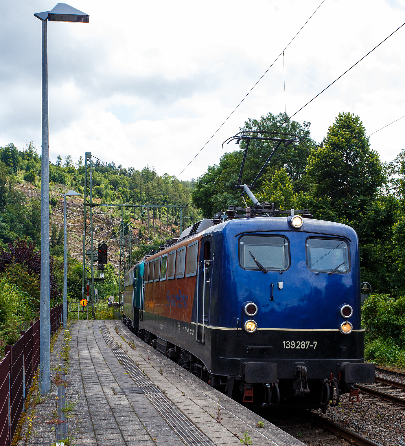 Die 139 287-7 (91 80 6139 287-7 D-BYB) mit der kalten 140 850-9 (91 80 6140 850-9 D-BYB) beide von der BayernBahn GmbH fahren am 04 Juli 2024 mit dem sogenannten  Henkelzug  (Langenfeld/Rhld. nach Gunzenhausen), durch den Bahnhof Kirchen (Sieg) in Richtung Siegen.

Die 139 287-7:
Vorne die 139 287-7 wurde 1963 von Krauss-Maffei AG in München-Allach unter der Fabriknummer 18956 gebaut, er elektrische Teil ist von den Siemens-Schuckert-Werke (SSW) in Berlin. Als DB E10 287 wurde in Dienst gesetzt, mit der Einführung des EDV-Nummernsystems wurde sie zum 01.01.1968 zur DB 110 287–0. Die Lok ist mit Einholmstromabnehmern ausgerüstet, sie ist die letzte gebaute Kasten 110er. Zur 139er wurde sie erst 1994 nach einem Umbau, der Lokkasten wurde auf Drehgestelle der Baureihe 140 (E40) gesetzt und es folgte die Umzeichnung in DB 139 287-7. Zum Mai 2009 erfolgte die Z-Stellung bei der DB AG. Zum 1.Oktober 2011 wurde die Lok durch die BayernBahn GmbH erworben und erhielt anschließend eine Untersuchung. Die Neulackierung in kobalt-blau/orange erhielt sie dann 2017.

Die 140 850-9
Die (E40) 140 850-9 wurde 1972 bei Henschel & Sohn in Kassel unter der Fabriknummer 31696 gebaut, der elektrische Teil wurde von der AEG in Berlin unter der Fabriknummer 8804 geliefert/gebaut. Als 140 850-9 wurde sie an die Deutsche Bundesbahn ausgeliefert, die Ausmusterung bei der DB Cargo AG erfolgte im Juli 2017 und sie wurde an die BayernBahn GmbH in Nördlingen verkauft.