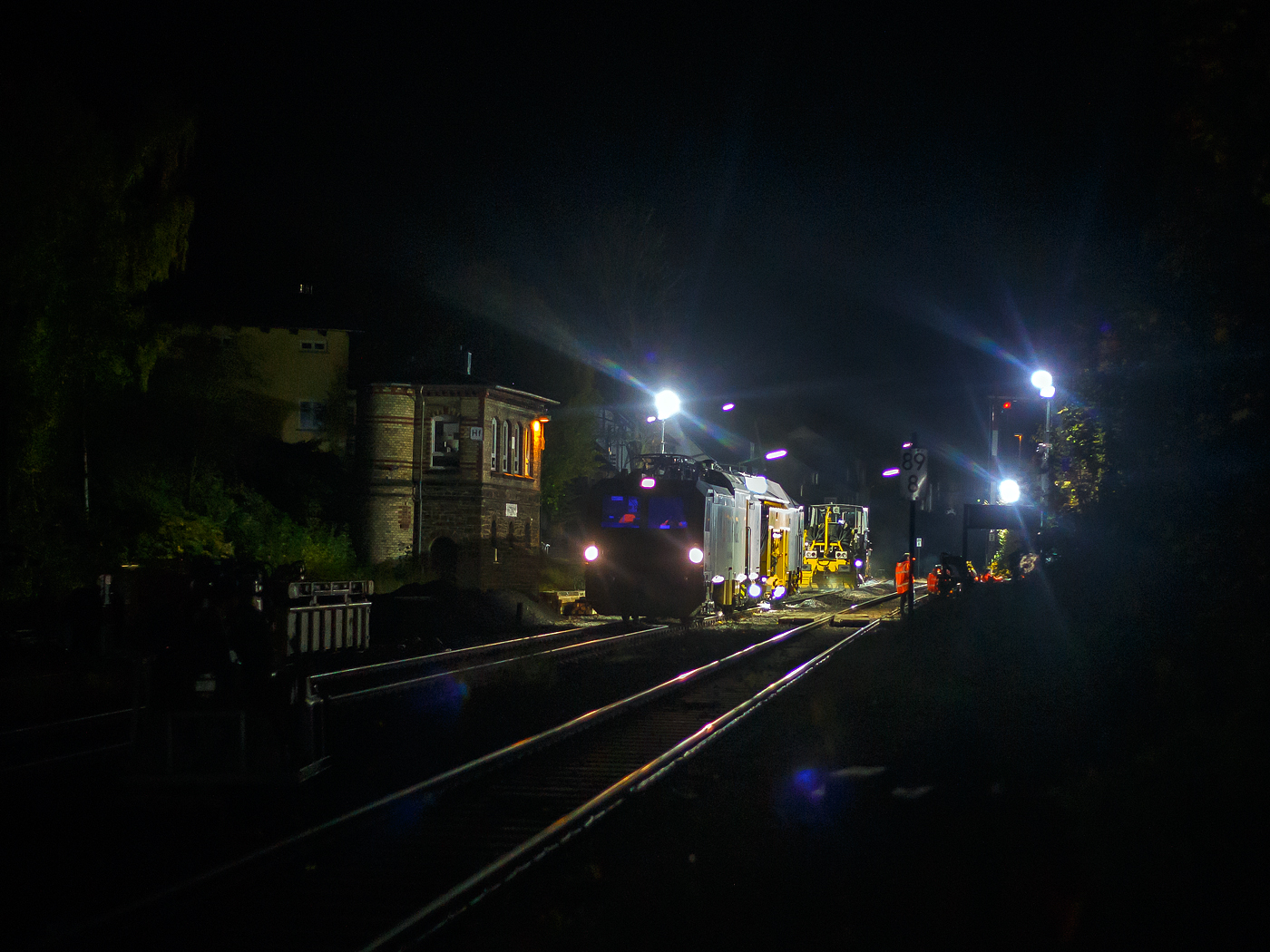 Die Baustelle Bahnhof Herdorf am Abend (19:15 Uhr) des 26 Oktober 2025, nach der Zeitumstellung ist es nun um diese Zeit wieder stockdunkel. Die Gleisbauer der KAF Falkenhahn Bau AG (Kreuztal) waren schon gegen 16 Uhr, noch bei Tageslicht, fertig mit dem Einschottern und mussten dann auf die Stopf- sowie die Schotterplaniermaschine warten. Nun sind beide angekommen. Ich hatte hier vom Bahnsteig im Bahnhof die Ankunft festgehalten und da ich schon öfter das Stopfen gesehen und dokumentiert habe, so ging es wieder nach Hause. 

Zu Hause angekommen habe ich die Bilder auf den PC geladen und diese bearbeitet, als ich dann sah, was ich fotografiert hatte, was ich Vorort nicht sehen konnte, so musste ich trotz der Dunkelheit nochmals zur Baustelle. Aber ich wählte dann, auf der anderen Seite beim Stellwerk Herdorf Fahrdienstleiter in der August-Sander-Straße aus. Übrigens die Straße ist nach dem 1876 in Herdorf geboren bekannten deutschen Fotograf August Sander benannt. Dieser Fotostandort war weitaus besser und näher am Geschehen, als hier vom Bahnhof.  