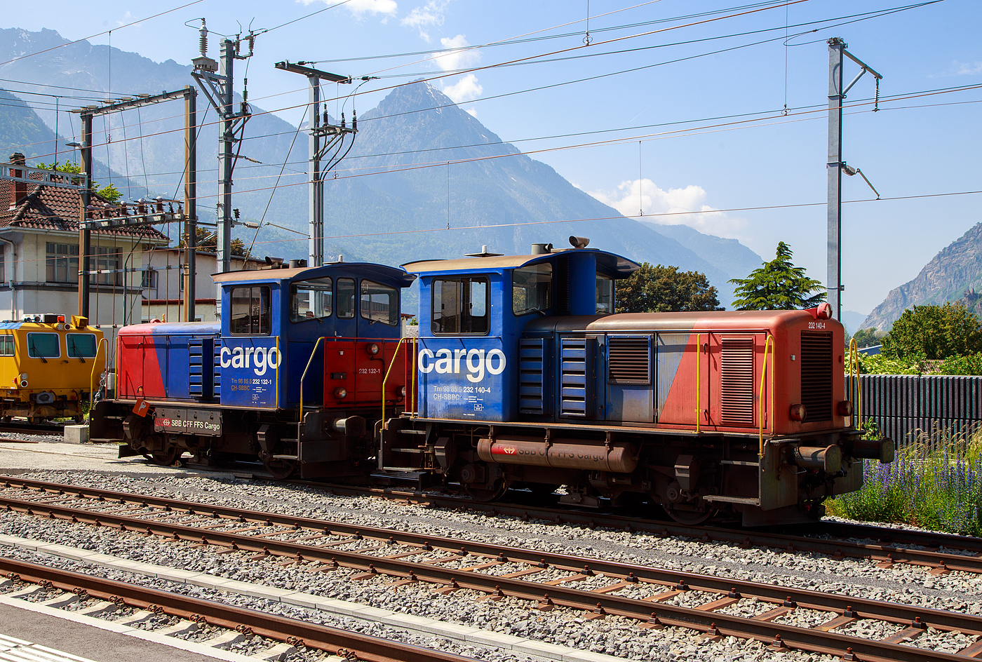 Die beiden ex SBB Tm IV Diesellok (Rangiertraktor mit Dieselantrieb), die 232 132-1 (Tm 98 85 5 232 132-1 CH-SBBC) und die 232 140-4 (Tm 98 85 5 232 140-4 CH-SBBC) sind am 26.05.2023 beim Bahnhof Martigny abgestellt.

Die Tm IV wurden 1975 von der SLM (Schweizerische Lokomotiv- und Maschinenfabrik) in Winterthur gebaut. Die 232 132-1unter der Fabriknummer 5048 als Tm 8782 und die 232 140-4 1unter der Fabriknummer 5048 als Tm 8790, jeweils f�r die SBB - Schweizerische Bundesbahnen, 2001 gingen sie an die SBB Cargo �ber. Beide wurden 2010 bzw. 2011 im Industriewerk Biel der SBB zu den heutigen Tm 232 umgebaut, es erfolgten eine Remotorisierung und der Einbau einer Funkfernsteuerung. Wobei normalerweise m�sste man sie mit der Funkfernsteuerung nun als Tmf f�hren.

Die Fahrzeuge wurden als fahrdrahtunabh�ngiges Fahrzeug f�r den Rangier- und Baudienst beschafft. Erstmals bei einer gro�en SBB-Fahrzeugserie, kam bei diesen Fahrzeugen die hydraulische Kraft�bertragung zum Einsatz. Da das Turbowendegetriebe eine Ausr�stung f�r beide Fahrtrichtungen hatte, konnte auf das st�rungsanf�llige mechanische Wendegetriebe verzichtet werden. Um die ung�nstige Wirkungsgradcharakteristik abzuschw�chen, wurde ein nur im Stillstand schaltbares Stufengetriebe eingebaut, das die Wahl zwischen einer H�chstgeschwindigkeit von 30 oder 60 km/h, oder die Wahl zwischen gr��erer Zugkraft oder gr��erer Geschwindigkeit erm�glichte.

Die Rangiertraktoren besitzen einen Au�enrahmen mit obenliegenden Blattfedern, die sich auf die Achslager abst�tzen. Die Federn besitzen keinen Lastausgleich.Die Kraft�bertragung erfolgt von der Motorwelle �ber eine Kardanwelle (Gelenkwelle) zu einem zentralen Schaltgetriebe. Dieses ist als Voith-Turbogetriebe mit nachgeschaltetem Verteilgetriebe ausgef�hrt. Von dessen Antriebswelle (beidseitig)  weiter �ber zwei Gelenkwellen auf eine Achsgetriebe, das sich auf der jeweiligen Antriebachse befindet. Die beiden Achsen sind �ber die Gelenkwellen und Antriebswellen mechanisch verbunden.

Das Voith-Turbogetriebe ist ein vollautomatisch arbeitendes hydrodynamisches Str�mungsgetriebe, das zwei Wandler besitzt. Als Betriebsfl�ssigkeit kommt Mineral�l zu Einsatz. Die beiden Wandler sind zueinander gegenl�ufig, wodurch die Fahrtrichtung ge�ndert wird. Dem Turbogetriebe ist das Verteilgetriebe nachgeschaltet. Dort kann mechanisch die �bersetzung zwischen Rangier- und Streckengang geschaltet werden. Da es sich um ein nicht synchronisiertes Schaltgetriebe handelt, darf es nur im Stillstand geschaltet werden. Es besitzt auch eine Neutralstellung, in welchen der Schalthebel bei Schleppfahrt gebracht und verriegelt wird. Das Achsgetriebe besteht aus einem Kegelradgetriebe mit einem �bersetzungsverh�ltnis 1:3.357. Auf der der Kardanwelle abgewandten Seite ist das Geh�use des Achsantriebes mit einer Drehmomentabst�tzung mit dem Fahrzeugrahmen verbunden.

Als Motor wurde ein schnelllaufender nicht aufgeladener 8-Zylinder Dieselmotor eingebaut. Die ersten Fahrzeuge waren mit einem MAN Motor des Typs R8V 16/18, mit 280 kW / 380 PS Leistung, ausger�stet, dessen Produktion sp�ter von der MTU �bernommen wurden. 

Umbau zu Tm 232
Neben der Neumotorisierung war der Einbau einer Zugsicherung der wichtigste Umbaupunkt, denn es werden doch heute vermehrt Streckenfahrten mit den Rangiertraktoren unternommen. Es kommt ein Caterpillar 6 Zylinder-Dieselmotor des Typs 13C Acert mit Ladeluftk�hler und Partikelfilter zum Einbau. Die Motorleistung betr�gt nun 310 kW, mehr l�sst das hydraulische Getriebe auch nicht zu. Daneben wurde die Spannung der Steuerung von 36 Volt auf 24 Volt abge�ndert. Neben dem Einbau einer neuen Sicherheitssteuerung und Zugsicherung werden auch alle Fahrzeuge mit einer modernen Funkfernsteuerung ausger�stet. Die Umbauten wurden im Industriewerk Biel durchgef�hrt, das heute der SBB Division Personenverkehr zugeteilt ist.

TECHNISCHE DATEN (Tm 232, ex Tm IV)
Spurweite:  1.435 mm (Normalspur)
Achsformel: B
L�nge �ber Puffer:  7.670 mm
Achsabstand:  3.570 mm
Treibraddurchmesser:   950 mm
Kleinster bef. Halbmesser:  35m
Dienstgewicht:  30 t
H�chstgeschwindigkeit: 30 (Rangiergang) / 60 km/h (Streckengang) / 90 km/h (geschleppt)
Traktionsleistung:  310 kW/421 PS (urspr. 280 kW / 380 PS)
Anfahrzugkraft: 90 kN
Dauerzugkraft:  60 kN
Motor (neu): 6 Zylinder Reihenmotor vom Typ Caterpillar 13C Acert)
Urspr�nglicher Motor:  8 Zylinder Reihenmotor vom Typ MAN Typs R8V 16/18
Leistungs�bertragung:  Hydraulisch
Tankinhalt:  850 l (Diesel)