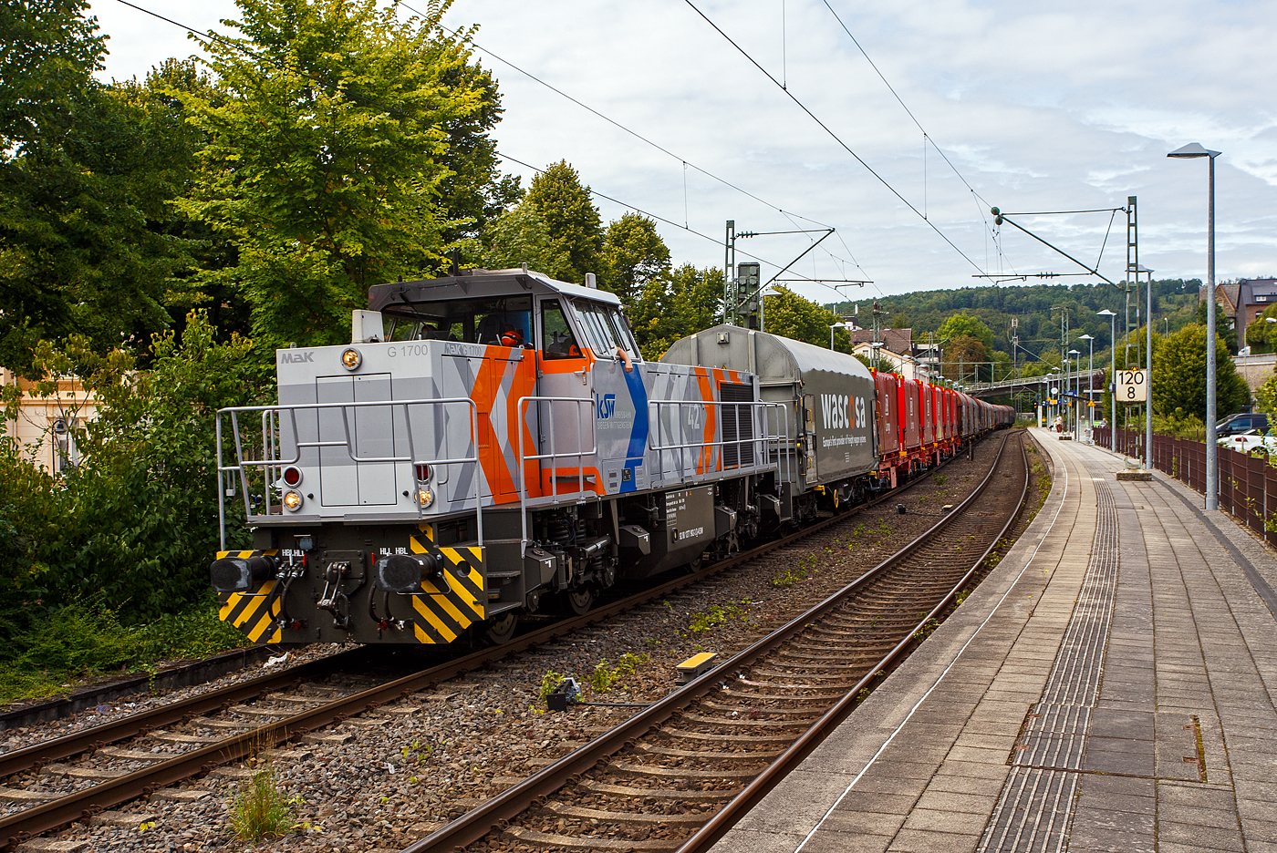 Die KSW 42 bzw. 277 902-3 (92 80 1277 902-3 D-KSW) der KSW (Kreisbahn Siegen-Wittgenstein), f�hrt am 22 August 2025, mit einem Coilzug durch Kirchen/Sieg in Richtung Betzdorf/Sieg. In Betzdorf setzt sie dann um und bringt dann den Coilzug, �ber die KBS 462 „Hellertalbahn“, nach Herdorf auf den KSW Rangierbahnhof Herdorf (Freien Grunder Eisenbahn KSW NE447 / DB-Nr. 9275). Nochmals einen lieben Gru� an das nette Lokpersonal zur�ck.

Es ist f�r die n�chsten 3 Wochen die letzte Fuhre �ber die KBS 460 „Siegstrecke“ nach Herdorf, da die Strecke zwischen Siegen und Troisdorf wegen Bauarbeiten dann gesperrt ist. Dann muss die KSW �ber die KBS 445 „Dillstecke“ bis Dillenburg fahren, nach dem Umsetzen dort geht es dann wieder ca. 5 km zur�ck nach Haiger und dann auf die „Hellertalbahn“ (Bahnstrecke Betzdorf–Haiger) nach Herdorf. Aber nicht nur der Umweg ist ein Nachteil, sondern der gr��te Nachteil ist die Streckenklasse CE (Radsatzlast 20,0 t / Meterlast 8,0 t/m) der „Hellertalbahn“ zwischen Haiger und Herdorf. Zwischen Herdorf und Betzdorf hat sie D4 (Radsatzlast 22,5 t / Meterlast 8,0 t/m). So k�nnen Coilwagen dann nicht mit voller Nutzlast verkehren, sondern m�ssen abgelastet werden, d.h. das sich z.B. nur 2 Coils anstelle von 3 Coils befinden auch wenn die 20 t Radsatzlast nicht ganz erreicht sind. 

Die Lok ist eine Vossloh MaK G 1700 BB sie wurde 2001 unter der Fabriknummer 1001108 bei Vossloh gebaut und wurde an die damalige SK - Siegener Kreisbahn GmbH (seit 2004 KSW) geliefert. Sie war die erste gebaute G 1700 BB.

Die Vossloh Lokomotive MaK G 1700 BB ist eine dieselhydraulische Lokomotive. Bei der �berarbeitung der Typenpalette nach der �bernahme der MaK durch Vossloh 1998 wurde im 4. Typenprgramm unter der Bezeichnung G 1700 BB der Bau einer vierachsigen Streckenlokomotive mit Mittelf�hrerhaus vorgesehen. Tats�chlich handelte sich zun�chst um eine Weiterentwicklung der G 1206. Die Maschine wurde in einigen Details an die Lokomotiven des 4. Typenprgramms angepasst. Dies betraf in erster Linie den Rahmen, der so ge�ndert wurde, dass die neuen Standarddrehgestelle verwendet werden konnten. Dabei wurden die Rahmenenden so umgestaltet, dass die Lokomotiven die im 4. Typenprgramm �blichen Komfortaufstiege erhielten. Ebenfalls ge�ndert wurde der Dieselmotor. Zum Einsatz kam der schon bei den G 1206 f�r die Dortmunder Eisenbahn verwendete MTU 12V 4000 R20 mit einer unver�nderten Leistung von 1500 kW. Die �brigen Komponenten der G 1700 BB blieben gegen�ber der G 1206 weitgehend unver�ndert.

Die ersten beiden Lokomotiven dieses Typs (die erste f�r die SK) wurden im Jahr 2001 ausgeliefert. Ende 2002 und Anfang 2003 folgten weitere vier Lokomotiven als Vorratsbauten. Nach diesen sechs Lokomotiven wurde der Bau der G 1700 BB zugunsten der technisch sehr �hnlichen und bereits weiter verbreiteten G 1206 wieder eingestellt.

Bei der G 1700-2 BB handelt es sich nicht um eine Weiteentwicklung der G 1700 BB, sondern um eine komplette Neuentwicklung. 

TECHNISCHE DATEN:
Spurweite: 1.435 mm (Normalspur)
Achsfolge: B´B´  
L�nge �ber Puffer: 14.700 mm 
Drehzapfenabstand: 7.400 mm 
Drehgestellachsstand: 2.400 mm 
gr��te Breite: 3.080 mm
gr��te H�he �ber Schienenoberkante: 4.220 mm
Raddurchmesser: 1.000 mm (neu)
kleinster befahrbarer Gleisbogen: 60 m
Dienstgewicht: 87,3 t 
Kraftstoffvorrat: 3.500 l
Motor: MTU-12-Zylinder-Dieselmotor, vom Typ 12V4000R20 
Leistung: 1.500 kW bei 1800 U/min
Getriebe: Voith L 5r4 zU2 
H�chstgeschwindigkeit: 100 km/h 
Baujahre: 2001 - 2003 
