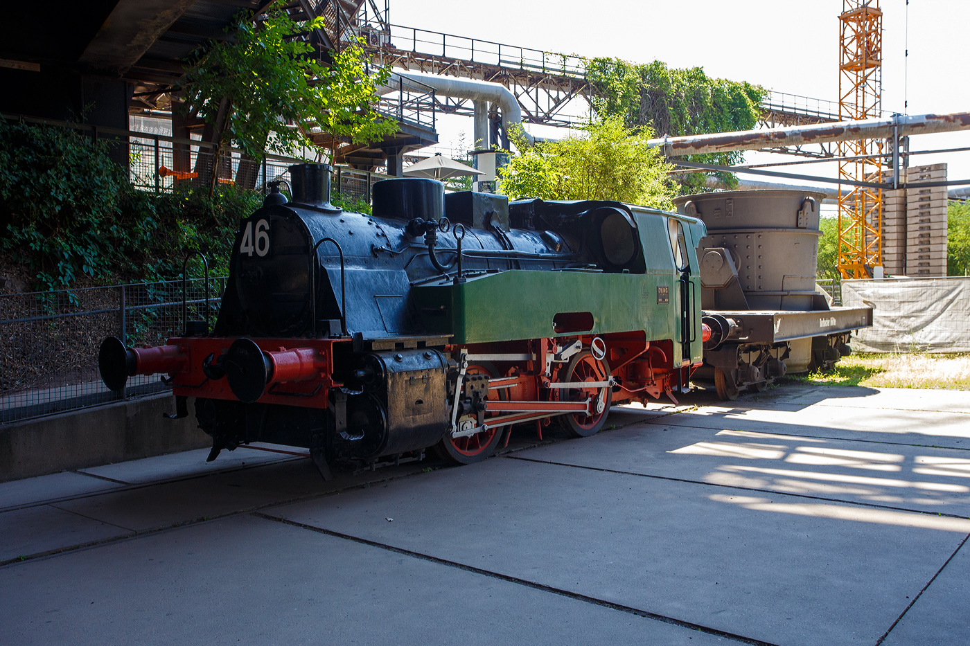 Die Normalspur-Dampflokomotive 46 der V�lklinger H�tte (ehem. R�chling'schen Eisen- und Stahlwerke GmbH in V�lklingen/Saar, ab 1982 ARBED-Saarstahl GmbH, ab 1986 Saarstahl V�lklingen GmbH), eine Jung Hochofenlok niedriger Bauart, abgestellt mit einem offenen Roheisenpfannenwagen der Burbacher H�tte, hier am 01 Juli 2025 am Gel�nde V�lklinger H�tte als Denkmal. Heutiger Eigent�mer ist die Initiative V�lklinger H�tte e.V..

1964 wurde die Lok Nr. 46 mit der Fabriknummer 13835 im Auftrag der R�chling'schen Eisen- und Stahlwerke als letzte rostgefeuerte Dampflok der Firma Arnold Jung, Lokomotivfabrik GmbH, in Jungenthal bei Kirchen an der Sieg gebaut und am 09.12.1964 geliefert. Einsatzgebiet war der Roheisentransport von den sechs Hoch�fen zum Thomasstahlwerk, wobei die besonders beengten Verh�ltnisse auf der V�lklinger H�tte eine besondere Bauform erforderten. Die Verbindung zwischen Eisenwerk und Stahlwerk war nur durch einen Tunnel m�glich, da die Deutsche Bahn durch ihre Gleisanlagen die H�tte in zwei Bereiche teilt. Dieser Tunnel, der Roheisenkanal, ist so niedrig, dass die zweiachsige Lok nur eine H�he von 2.640 mm �ber der Schienenoberkante und eine Breite von 2.450 mm aufweist. Die Loks 44 (11945/Baujahr 1953) und 45 (13240/Baujahr 1959), die nach vorliegenden Unterlagen technisch baugleich sind, wurden schon vorher (von der Arnold Jung GmbH an die V�lklinger H�tte geliefert. 

Um speziell die F�hrerhausr�ckwand, die Lampen, aber auch die Kupplungselemente zwischen Lok und dem ersten Wagen vor fl�ssigen gl�henden Spritzern von herabfallenden Roheisen zu sch�tzen, erhielten die Lokomotiven ein nach hinten verl�ngertes Dach. Die Puffer wurden im Stahlwerk den dort vorhanden Roheisenpfannenwagen angepasst. Alte Jung Mitarbeiter berichten, an der Lok h�tte man kaum was verdient, weil l�nst nicht mehr Dampflokteile ab Lager vorhanden waren umd z.T. m�hsam und teuer beschafft werden musste.

Im Zuge der Modernisierung und Rationalisierung wurden die Roheisendampfloks 1981 nicht mehr ben�tigt. W�hrend die Lokomotiven 44 und 45 an ein Museum im Ruhrgebiet verkauft wurden, gelangte die Lokomotive 46 als technisches Denkmal 1982 zur Universit�t Kaiserslautern. Diese schenkte die Lokomotive 46 der Initiative V�lklinger H�tte, die sie mit Hilfe von Sponsoren am 9. Februar 1999 nach V�lklingen zur�ckholte.

TECHNISCHE DATEN:
Baujahre: 1953,1959 und 1964
Gebaute Anzahl: 3
Spurweite: 	1.435 mm (Normalspur)
Achsformel: B
L�nge: 7.600 mm
Achsabstand: 2.000 mm
Triebraddurchmesser: 1.000 mm (neu)
Breite: 2.450 mm
H�he: 2.650 mm
Dienstgewicht: 26 t
H�chstgeschwindigkeit: k. A. 
Leistung: 250 PS
Zugkraft: 7,5 t
Zylinder: 2 St�ck, � 380 mm x 500 mm Hub
Dampfdruck: 14 bar
Heizfl�che: 58,92 m�
Rostfl�che: 1,17 m�
Anzahl Siederohre: 178 St�ck
Durchmesser Siederohre: 33/38 mm
L�nge Siederohre: 2.540 mm
Wasservorrat: 2.500 l (2,5 m�)
Kohlenvorrat: 500 kg