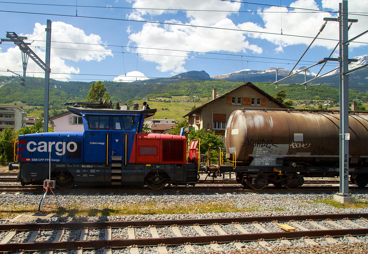 Die SBB Cargo Eem 923 002-0  Brästenegg  (Eem 97 85 1 923 007-9 CH-SBB C) rangieret, am 26.05.2023 beim Bahnhof Siders/Sierre (Kanton Wallis), einen Kesselwagen (aus dem Zug heraus aufgenommen).

Die Zweifrequenz-Hybridlokomotive vom Typ BUTLER wurde 2012 von Stadler Winterthur unter der Fabriknummer L-11000/002 gebaut. 

SBB Cargo bestellte im Juni 2010 beim Schweizer Fahrzeugbauer Stadler Rail in Winterthur 30 neue Zweifrequenz-Hybridlokomotive Typ Eem 923 001-030 für Güter- und Rangierdienste. Diese Hybridlok ist eine richtige Kompaktlok die unter Fahrleitungsbetrieb 15kV 16,7 Hz sowie 25kV 50 Hz Wechselstrom fahren kann und für die  letzte Meile  dank eines Dieselmotors auch auf nicht elektrifizierten Gleisen ohne Leistungsverminderung ihre Arbeit verrichten kann. Mit einer Leistung von 1.500 kW, einer Höchstgeschwindigkeit von 120 Km/h sowie Vielfachsteuerungsmöglichkeit für bis zu 4 Eem 923 ist diese Lok für SBB Cargo ein wahrer Alleskönner. 

Weitere Technische Merkmale sind:
Verschleiß lose elektrische Bremse mit Rückführung der Bremsenergie ins Fahrleitungsnetz (Rekuperation) oder über Bremswiderstände im Dieselbetrieb.
Pneumatische indirekte Bremse für Zug und direkte Bremse für die Lok.
Ein leistungsstarker Schraubenkompressor mit einer Förderleistung
von 2.400 Liter/min.

TECHNISCHE DATEN:
Hersteller: Stadler Rail Winterthur
Eigentümer: SBB Cargo
Anzahl der Fahrzeuge:  30
Spurweite: 	1.435 mm (Normalspur)
Achsfolge:  Bo
Länge über Puffer:  9.100 mm
Achsabstand:  4.300 mm
Fahrzeugbreite:  3.100 mm
Fahrzeughöhe:  4.306 mm
Dienstgewicht (Tara)  45 t
Raddurchmesser Neu:  1.100 mm
Maximale Leistung am Rad:  1.500 kW (2.040 PS)
Maximale Leistung Dieselmotor:  290 kW (394 PS)
Anfahrzugkraft: 150 kN
Höchstgeschwindigkeit:  120 km/h
Umgrenzungsprofil: UIC 505-I AB-EBV U3
Fahrdrahtspannung:  15kV 16,7 Hz und 25kV 50 Hz Wechselstrom
Zugsicherungsystem:  Signum-ZUB 262ct, ETM-S
Nachrüstungsmöglichkeiten:  ETCS-Level 2 / Indusi
Zugfunksystem:  GSM-R
Auslieferungsjahr:  2012-2013
