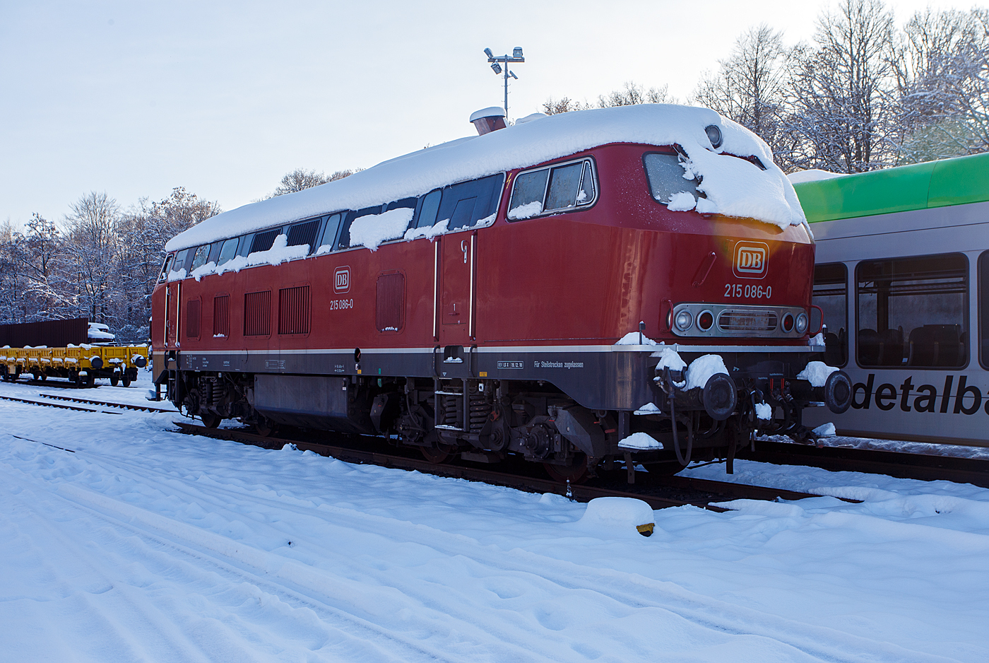 Nicht nur im Süden der Republik hat es etwas geschneit, wie hier auf dem Westerwald auf der Bindweide, aber nicht so mächtig.  Die für Steilstrecken zugelassenen 215 086-0 (eigentlich 225 086-8 / 92 80 1225 086-8 D-EVG) der RE - Rheinische Eisenbahn ist am ersten Advent  dem 3 Dezember 2023 auf dem Betriebshof der Westerwaldbahn (WEBA) auf der Bindweide bei Steinebach/Sieg abgestellt. Dahinter stehen noch die beiden Daadetalbahn STADLER GTW 2/6 VT 117 und VT 118 (646 417-5 und 646 418-3) der Westerwaldbahn (WEBA), ex VT 117 und ex VT 118 HellertalBahn, die beiden gingen nach Auflösung der HellertalBahn an die WEBA, der 3. der VT 116 ging an die HLB.

Die V 163 wurde 1971 bei MaK unter der Fabriknummer 2000091 und an die DB als 215 086-0 mit stärkerem Motor (1.840 kW) ausgeliefert und war bis 2000 bei der DB Regio AG. Im Jahr ging sie an die DB Cargo AG und wurde in die heutige 225 086-8 umgebaut und um gezeichnet.  Zum Umbau gehörte der Ausbau der Dampfheizung, da diese für Güterzüge nicht mehr benötigt wurde. Stattdessen wurde ein Vorwärm- und Warmhaltegerät für die Maschinenanlage eingebaut. Außerdem besitzen die Loks eine Doppeltraktionssteuerung, die Doppeltraktionen mit Loks der Baureihen 217 und 218 ermöglichen. 

Mit der Ausmusterung bei der nunmehrigen DB Schenker Rail Deutschland AG im Jahr 2011 erfolgte auch der Verkauf an die RE - Rheinische Eisenbahn, hier wurde sie wieder in den Auslieferzustand zurückversetzt und trägt nun die ursprüngliche Bezeichnung 215 086-0, ist aber eine 225er und hat so die NVR-Nummer 92 80 1225 086-8 D-EVG).

Die Lokomotiven der Baureihe 215 wurden kurzfristig als Variante der V160-Familie für den Personen- und Güterverkehr beschafft und dabei im Gegensatz zu anderen Baumustern der V160-Familie mit Heizdampferzeugern ausgerüstet. Geplant war sie im alten Baureihenschema als V 163. Diese Lokomotiven sollten später in die Baureihe 218 umgebaut werden können. Dazu erhielt sie bereits den um 400 Millimeter verlängerten Lokkasten anderer bis dahin nur als Prototyp vorhandener Baureihen. Der Hauptgrund für den Bau der Baureihe 215 lag darin, dass die Reihe 218 noch nicht Serienreife erreicht hatte, die Bundesbahn aber die restlichen noch vorhandenen Dampflokomotiven ersetzen wollte. 1968 bis 1970 wurden insgesamt 150 Lokomotiven gebaut.

Mitte der 1980er erhielten die Maschinen mit 12-Zylinder-V-Motor Abgashutzen auf dem Dach (wie diese). Sie bewirkten eine Beschleunigung und Umlenkung der Abgase, so dass bei klimatisierten Reisezugwagen, welche auf dem Dach die Luftansaugöffnungen hatten, eine Belästigung der Fahrgäste durch Abgase oder Abgasgerüche vermieden wurde. Weiterhin sorgten die Hutzen noch für andere Effekte: die bis zu 280 °C heißen Abgase wurden V-förmig an der Oberleitung vorbeigeleitet, so dass die Oberleitung nicht verrußt wurde und sich nicht zu sehr ausdehnte. Außerdem wurde die Gefahr von Böschungsbränden durch herabsinkende, heiße Rußpartikel reduziert. Auch eine Türschließanlage und Bremsgestängesteller zum automatischen Nachstellen des Bremskolbenhubes wurden nachgerüstet.

TECHNISCH DATEN:
Gebaute Stückzahl: 150
Spurweite:  1.435 mm (Normalspur)
Achsformel:  B'B'
Länge:  16.400 mm
Drehzapfenabstand:  8.600 mm
Achsabstand im Drehgestell:  2.800 m
Gesamtradstand: 11.400 mm
Gewicht: 79 Tonnen
Radsatzfahrmasse:  20,0 Tonnen
Höchstgeschwindigkeit: 140 km/h (90 km/h im Rangiergang) Motorentyp: MTU MA 12 V 956 TB 10 (ein V12-Zylinder-Diesel-Motor)
Dauerleistung: 1.840 kW (2.500 PS)
Nenndrehzahl: 1.500 min-1
Leistungsübertragung: hydraulisch, über Kardanwellen vom mittig angeordneten Getriebe zu den beiden zweiachsigen Drehgestellen. So werden über Radsatzgetriebe jeweils beide und somit alle Achsen angetrieben. Ein Wendegetriebe ermöglicht den Richtungswechsel.
Getriebe: Voith L 820 brs  
Tankinhalt: 3.050 l (Kraftstoff) 
Sandvorrad: 320 kg 
Sie ist für Steilstrecken zugelassen.
