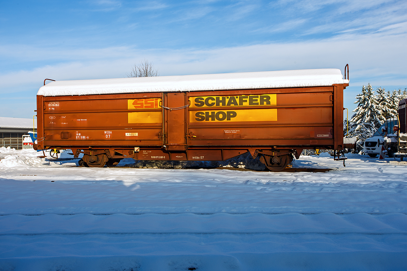 Nicht nur im Süden der Republik hat es etwas geschneit, wie hier auf dem Westerwald auf der Bindweide, aber nicht so mächtig. Der private gedeckte zweiachsige Güterwagen mit Schiebewänden und Schiebedach WEBA 07 - 10 80 0715 001-5 D-WEBA (ex 10 80 071 5 001-5 D-DB) der Gattung Tbis 869 - Bauform A (für Büromöbel der SSI Schaefer Shop GmbH) der Westerwald WEBA abgestellt am 03 Dezember 2023 (am 1. Advent) auf dem Betriebshof der Westerwaldbahn (WEBA) auf der Bindweide bei Steinebach/Sieg. Der Wagen ist auf DB-Strecke Scheuerfeld – Betzdorf(Sieg) zugelassen. 

Beschreibung:
Ab 1966 wurden von der DB bei der Waggonunion Siegen (eigentlich Dreis-Tiefenbach, zuvor Siegener Eisenbahnbedarf A.G.) erste Wagen der Gattung Tbis 869 der Bauform A beschafft. Bei dieser Bauform konnte das Dach nur von einer kleinen oben liegenden Bühne an den Stirnwänden geöffnet werden. Zwei Jahre später wurde die Bauform B dieser Gattung gefertigt. Hierbei war es nun möglich das Dach mittels Handräder an einer Stirnseite der Waggons zu öffnen. Es war nun nicht mehr nötig auf das Dach zu steigen und verminderte somit auch die Unfallgefahr.

Eingesetzt wurden diese Waggons für den Transport von großen nässeempfindlichen Stückgütern (bei diesem Wagen waren es Büromöbel), durch das öffnungsfähige Dach könnte die Ladung auch mittels Kranen schnell be- und entladen werden. Von den ca. 2000 beschafften Fahrzeugen gelangten noch gut 1400 zur DB AG, dort wurden sie aber zügig ausgemustert und durch neuere Bauarten ersetzt. 

TECHNISCHE DATEN:
Spurweite: 1.435 mm (Normalspur)
Anzahl der Achsen: 2
Länge über Puffer :14.020 mm
Achsabstand: 8.000 mm
Eigengewicht: 18.500 kg
Ladelänge:12.774 mm
Ladebreite: 2.670 mm
Ladefläche: 33,0 m²
Rauminhalt: 85,5 m³
Seitenwand-Türöffnung (B × H): 5.660 × 2.100 mm
Lichte Öffnung bei geöffnetem Dach: 6.000 × 2.530 mm
Fußbodenhöhe über SO: 1.200 mm
Höchstgeschwindigkeit: 100  km/h 
Maximales Ladegewicht: 18,5 t (Streckenklasse C oder höher)
Kleinster bef. Gleisbogenradius:  35 m
Bremse: Frein O-GP 
Intern. Verwendungsfähigkeit: RIV (ursprünglich)
