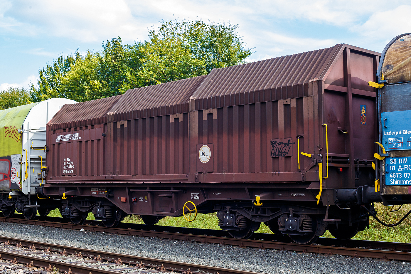 Vierachsiger Drehgestell-Flachwagen mit verschiebbaren Teleskophauben f�r Coiltransporte, 31 81 4668 333-5 A-RCW, der Gattung Shimmns, der Rail Cargo Wagon - Austria GmbH (ein Unternehmen der �BB), abgestellt im Zugverband (�bergabeg�terzug) am 15 September 2024 in Herdorf auf dem Rbf der Kreisbahn Siegen-Wittgenstein (KSW).

Der Wagen wurde 1985 von den Jenbacher Werke AG in Jenbach (�sterreich) unter der Fabriknummer 3.931-334 gebaut.

Der Wagen eignet sich speziell f�r den Transport witterungsempfindlicher Blechrollen. Er besitzt feste Stirnw�nde, 5 fest im Untergestell eingebaute Lademulden und drei teleskopartig angeordnete Hauben, die sich zu beiden Wagenenden hin soweit durch Ineinanderschieben �ffnen lassen, dass jeweils 2/3 des Wagens freigelegt werden kann. Hierdurch ist eine einfache Beladung des Wagens sowohl von oben als auch von der Seite aus m�glich. Der Wagen darf nur mit geschlossenen und verriegelten Hauben verkehren.

Das Untergestell und die festen Stirnw�nde, die als Abkantrahmenkonstruktion mit Blechbekleidung und hutf�rmigen Verst�rkungsprofilen ausgef�hrt sind, sowie die fest auf den Au�enlangtr�gern angeordneten drei Laufschienen je Wagenseite bilden den als Schwei�konstruktion ausgef�hrten Wagenkasten. Das Untergestell mit seinen beiden kr�ftigen Au�enlangtr�gern besteht aus Walz- und Abkantprofilen. Die Untergestellenden bestehen aus den Hauptquertr�gern, den AK-Tr�gern und den Kopfst�cken. Die sattelf�rmig angeordneten, jeweils mit den Au�enlangtr�gern verschwei�ten Quertr�ger bilden zusammen mit 6 mm dicken Muldenblechen die Lademulden.

Die teleskopartig ineinander verschiebbaren Stahlhauben bestehen aus einer Hohlprofil-Rahmenkonstruktion und einer mit Sicken versehenen Beblechung. Die Abdichtung der Hauben untereinander und zu den Stirnw�nden hin erfolgt �ber labyrinthartig ausgef�hrten Umrandungsprofilen. Die Endhauben verriegeln in geschlossener Stellung automatisch. Das Entriegeln der Hauben ist sowohl vom Erdboden wie auch von der Rampe m�glich. Die beidseitig an jeder Stirnwand angeordneten Verriegelungseinrichtungen sind durch Wellen miteinander verbunden. Hierdurch ist das Entriegeln von nur einer Wagenseite aus m�glich. Jede Haube l�uft auf 4 jeweils und mit Abhebesicherungen versehenen Laufwagen mit nadelgelagerten Doppelrollen.

TECHNISCHE DATEN:
Spurweite: 1.435 mm (Normalspur)
Achsanzahl: 4 (in 2 Drehgestelle)
L�nge �ber Puffer: 12.040 mm
L�nge der Ladefl�che: 10.800 mm
Ladebreite (Mulden): 2.400 mm
Drehzapfenabstand: 7.000 mm
Achsabstand im Drehgestell: 1.800 mm
Laufraddurchmesser: 920 mm (neu)
Eigengewicht:  23.000 kg
H�chstgeschwindigkeit: 100  km/h (beladen) / 120 km/h (leer)
Maximales Ladegewicht: 67,0 t (ab Streckenklasse D4) / DB Netz 61,0 t ab CM3
Kleinster bef. Gleisbogenradius: 35 m
Bauart der Bremse: O-GP-A max. 58 t (LL)
Bremssohle: IB 116
Handbremse: Ja
Intern. Verwendungsf�higkeit: RIV