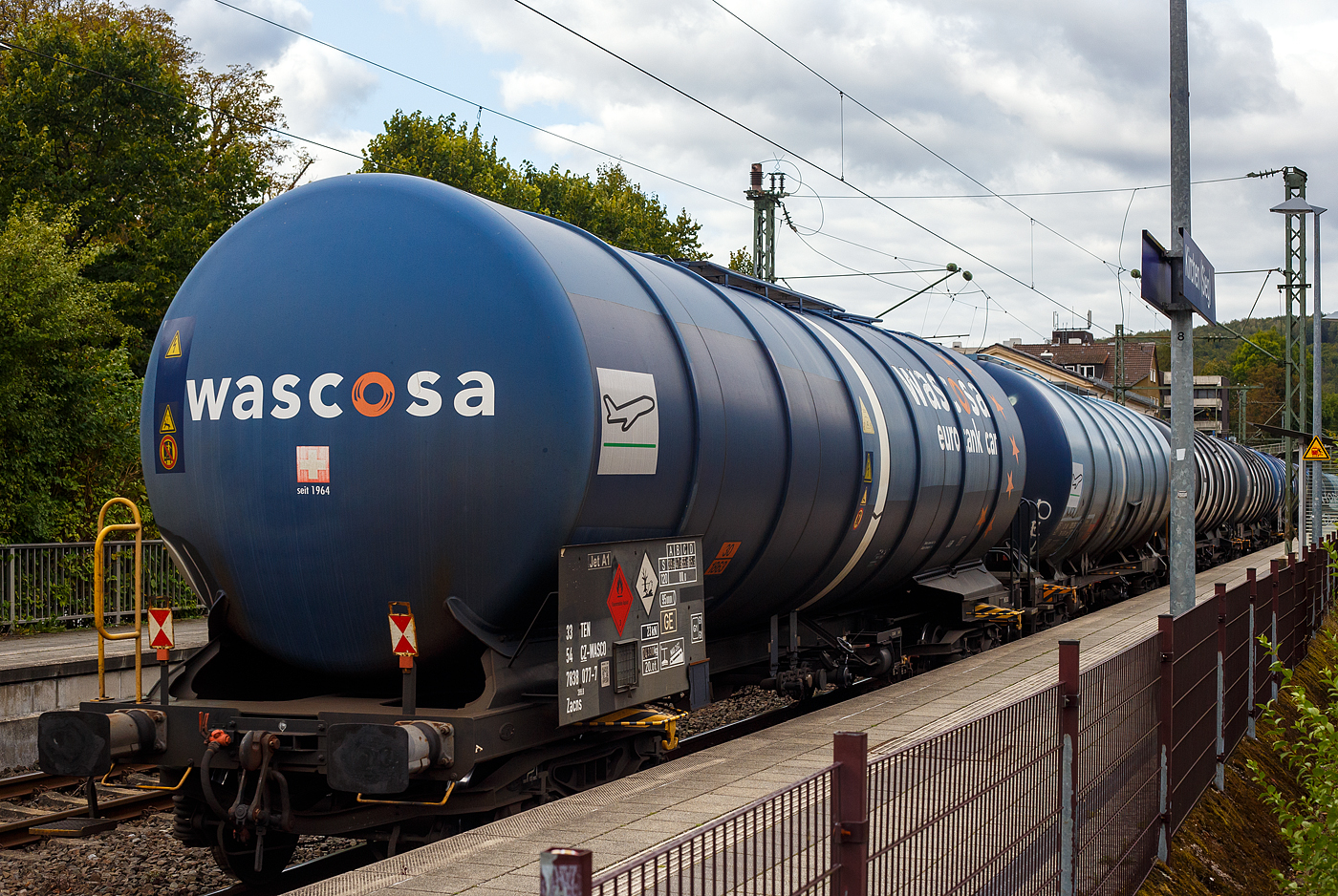 Vierachsiger großvolumiger Wascosa – euro tank car ® 95 m³-Drehgestell-Kesselwagen mit Bremserbühne (95 m³ Mineralöl-Kesselwagen) 33 54 7838 077-7 CZ-WASCO der Gattung Zacns 399.8, der Wascosa AG Luzern (CH) eigestellt in Tschechien, laut Gefahrguttafel/Warntafel 30/1863 beladen mit Düsenkraftstoff (auch bekannt als Kerosin bzw. leichtes Petroleum) hier laut Anschrift mit der Sorte Jet A-1 (NATO Code F-35), am 14 September 2024 im Zugverband (Zugschluss) bei der Zugdurchfahrt in Kirchen (Sieg).

Die Kesselwagen Wascosa – euro tank car ® , Zacns sind für den Transport von Mineralöl-Produkte, als optionale Sicherheitsausstattung besitzen sie Entgleisungsdetektor.

TECHNISCHE DATEN:
Spurweite: 1.435 mm (Normalspur)
Achsanzahl: 4 (in 2 Drehgestelle)
Länge über Puffer: 17.000 mm
Drehzapfenabstand: 11.460 mm
Drehgestelle: Y25 Lsd1(BA 628) , Radsätze BA 004
Achsabstand im Drehgestell: 1.800 mm
Laufraddurchmesser:  920 mm (neu) / 840 mm (abgenutzt)
Eigengewicht: 24.330 kg
Tankinhalt: 95.000 l
Max. Ladegewicht: 65,6 t (Streckenklasse D) 
Höchstgeschwindigkeit: 100 km/h (beladen) / 120 km (leer)
Kleinster befahrbarer Gleisbogen: R 35 m
Bremse: KNORR KE-GP (K)
Bremssohle: Cosid 810 BG 
Handbremse: 20 t
Intern. Verwendungsfähigkeit: TEN GE / G1

Tankcode: L 4 BH
Zul. Betriebsüberdruck: 3,0 bar
L= Tank für Stoffe in flüssigem Zustand (flüssige Stoffe oder feste Stoffe, die in geschmolzenem Zustand zur Beförderung aufgegeben werden)
4= zutreffender Mindestprüfdruck in bar
B = Tank mit Bodenöffnungen mit 3 Verschlüssen für das Befüllen oder Entleeren 
H = luftdicht verschlossener Tank
Werkstoff Tank: Feinkornbaustahl P355N (StE 355)

Das Transportgut Kerosin / Jet A-1 (NATO Code F-35):
Heute wird in der internationalen zivilen Luftfahrt mit Ausnahme der USA fast ausschließlich die Spezifikation Jet A-1 (entspricht der militärischen Bezeichnung JP-1A) mit etwas niedrigerem Gefrierpunkt (−47 °C), aber identischem Flammpunkt und Siedebereich wie Jet A als Flugturbinenkraftstoff verwendet. Der NATO-Code ist F-35. Jet A-1 hat eine Dichte zwischen 0,775 bis 0,825 kg/dm³, einen Flammpunkt von +38 °C und einen Gefrierpunkt von −47 °C.

Quellen: Anschriften und Datenblatt WASCOSA
